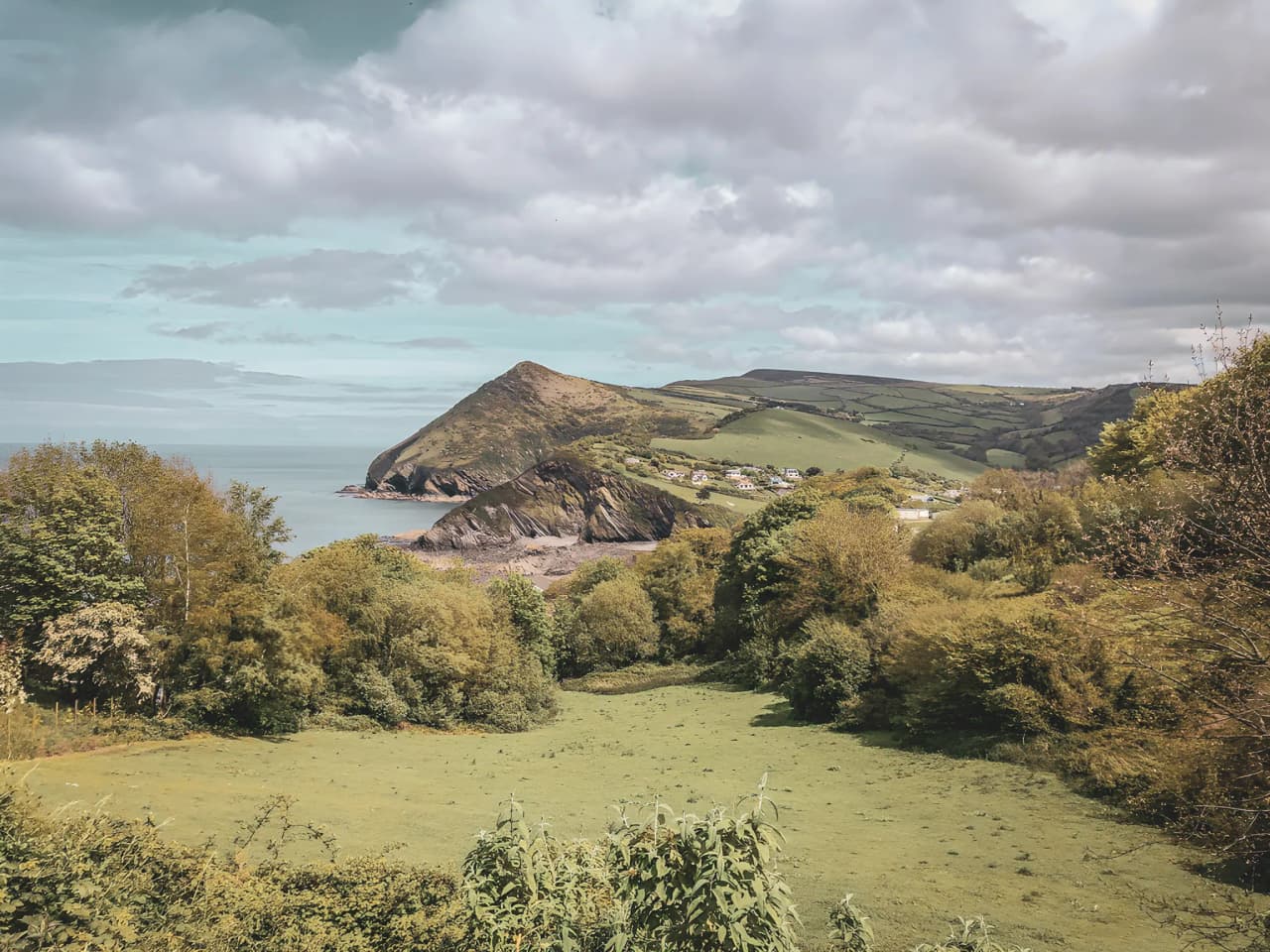 Scène côtière galloise avec des collines verdoyantes et une mer paisible sous un ciel nuageux.