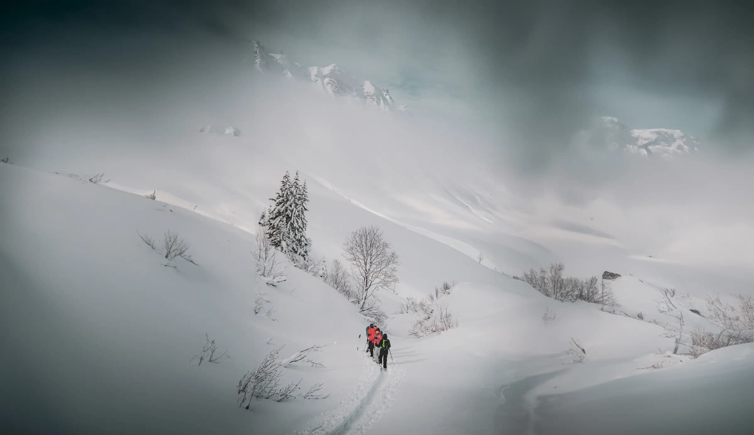 A snowy landscape with mountains in the background, where two hikers are making their way along a path through the snow. The hikers are wearing colourful jackets, contrasting with the dazzling white of the snow.