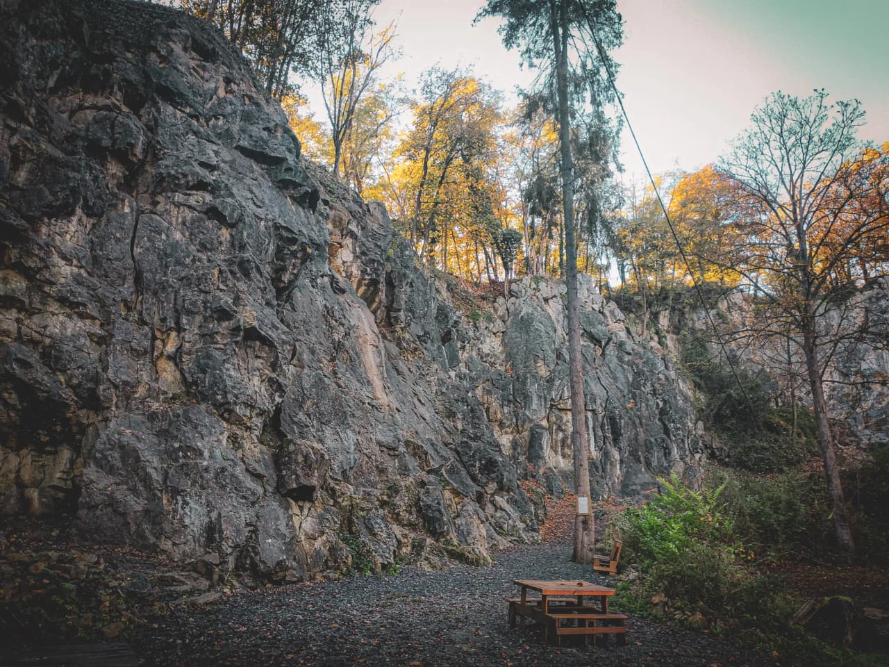 Un paysage de rocher escarpé, bordé d'arbres aux feuilles dorées qui signalent l'automne. Au premier plan, une table en bois avec des chaises est disposée sur un sol rocheux, entourée de