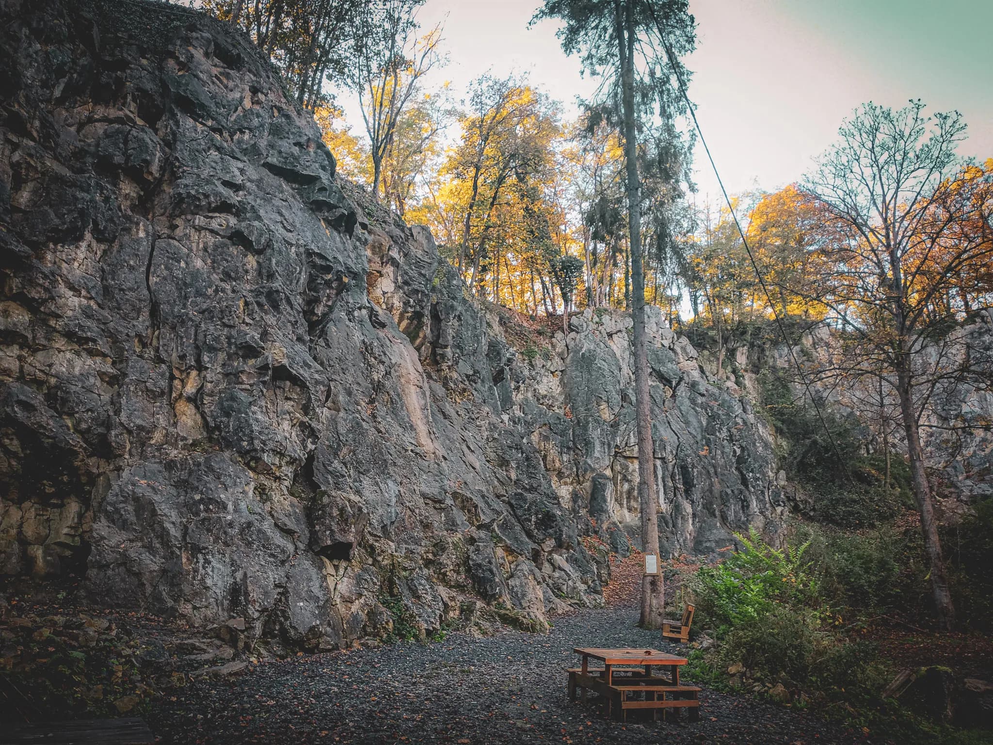 A landscape of craggy rock, bordered by trees with golden leaves that signal autumn. In the foreground, a wooden table with chairs is laid out on a rocky floor, surrounded by