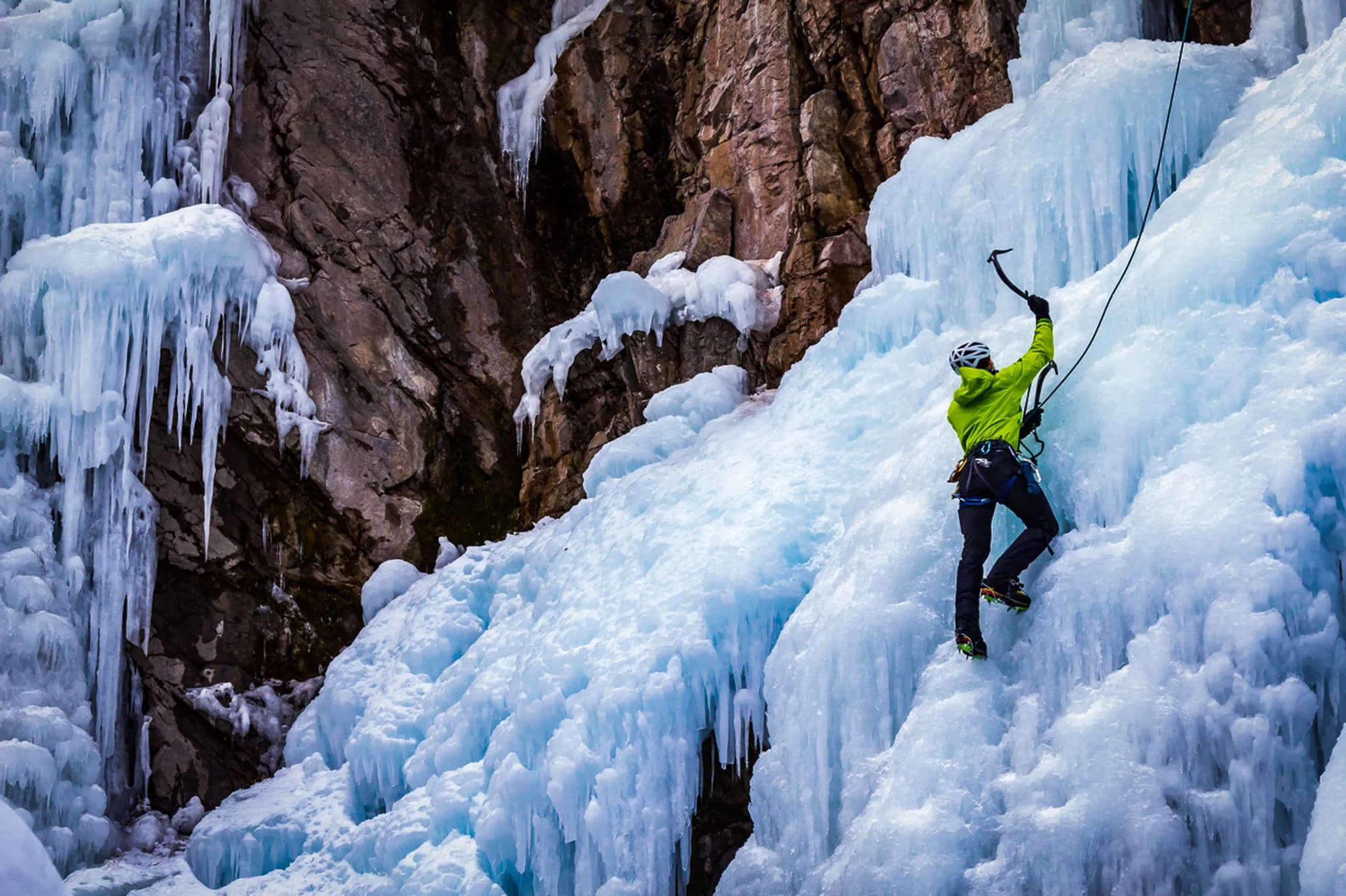 Un grimpeur en ascension sur un mur de glace, vêtu d'un manteau vert vif et d'un casque blanc, utilise un piolet pour progresser. Le relief est composé de formations de glace bleue et blanche qui s'acc