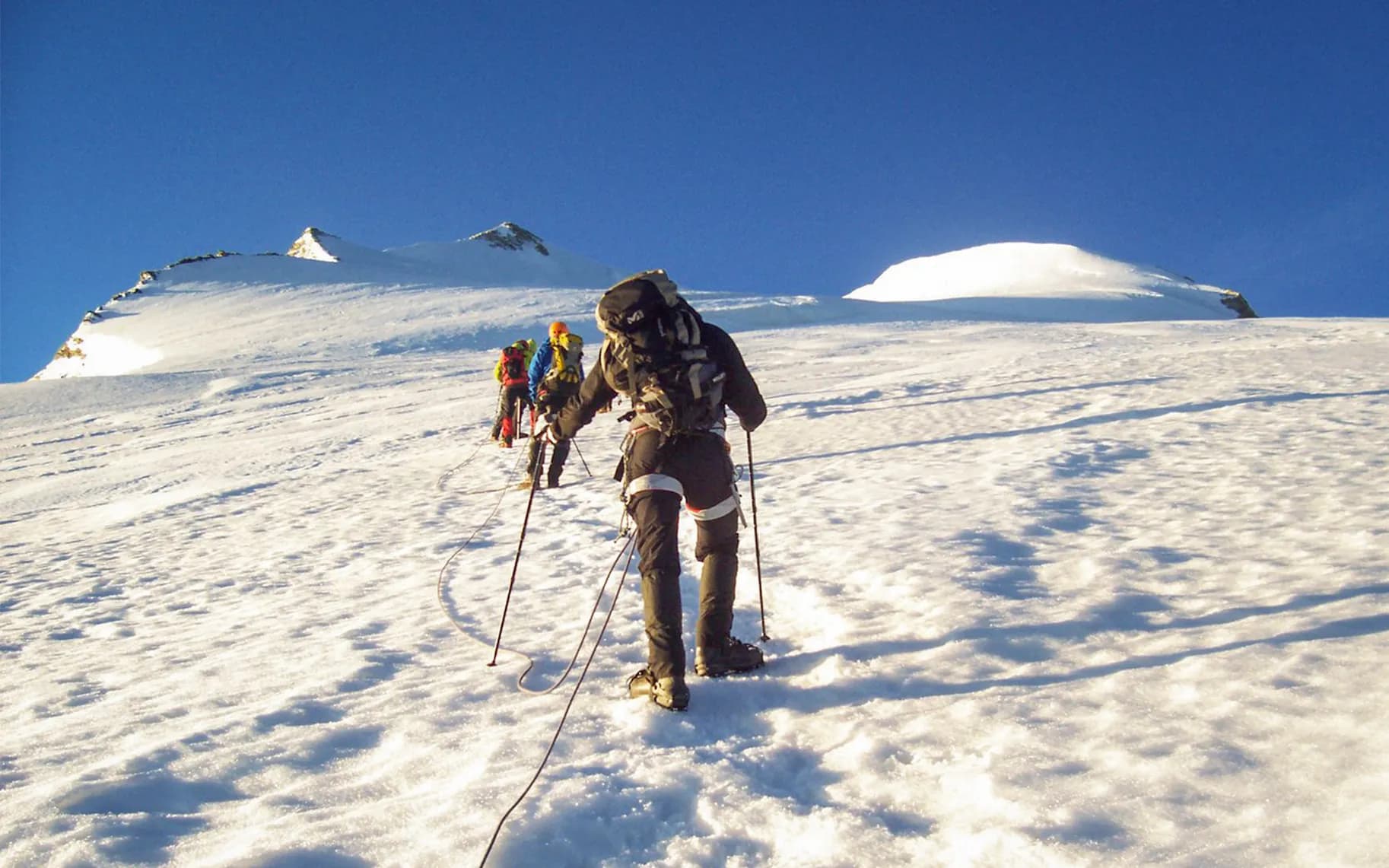 A group of mountaineers climb a snow-covered slope under a clear blue sky. The climbers are wearing protective clothing and are attached by a rope, advancing cautiously on the snow.