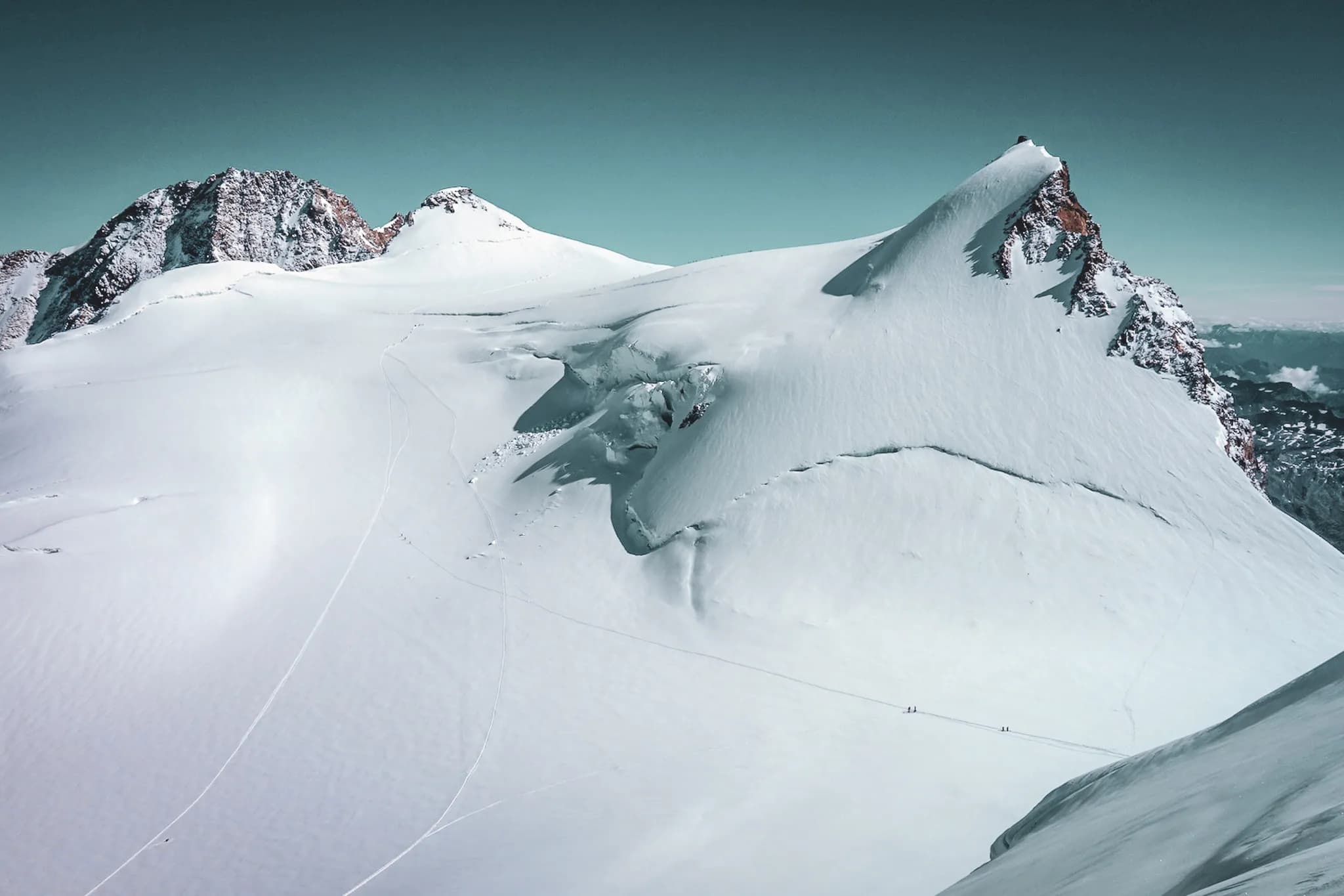 Een spectaculair Alpenlandschap gedomineerd door besneeuwde toppen. De grond is bedekt met een dikke laag smetteloze sneeuw, waardoor zachte, golvende vormen ontstaan. Op de achtergrond,