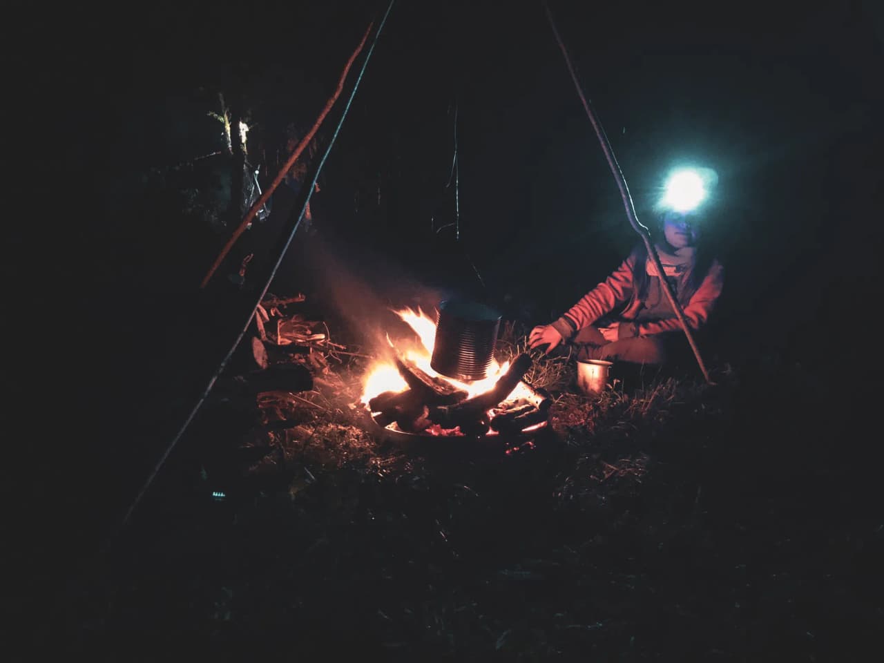 Un campeur auprès d'un feu de camp, éclairé par une lampe frontale, dans la nuit ardennaise.