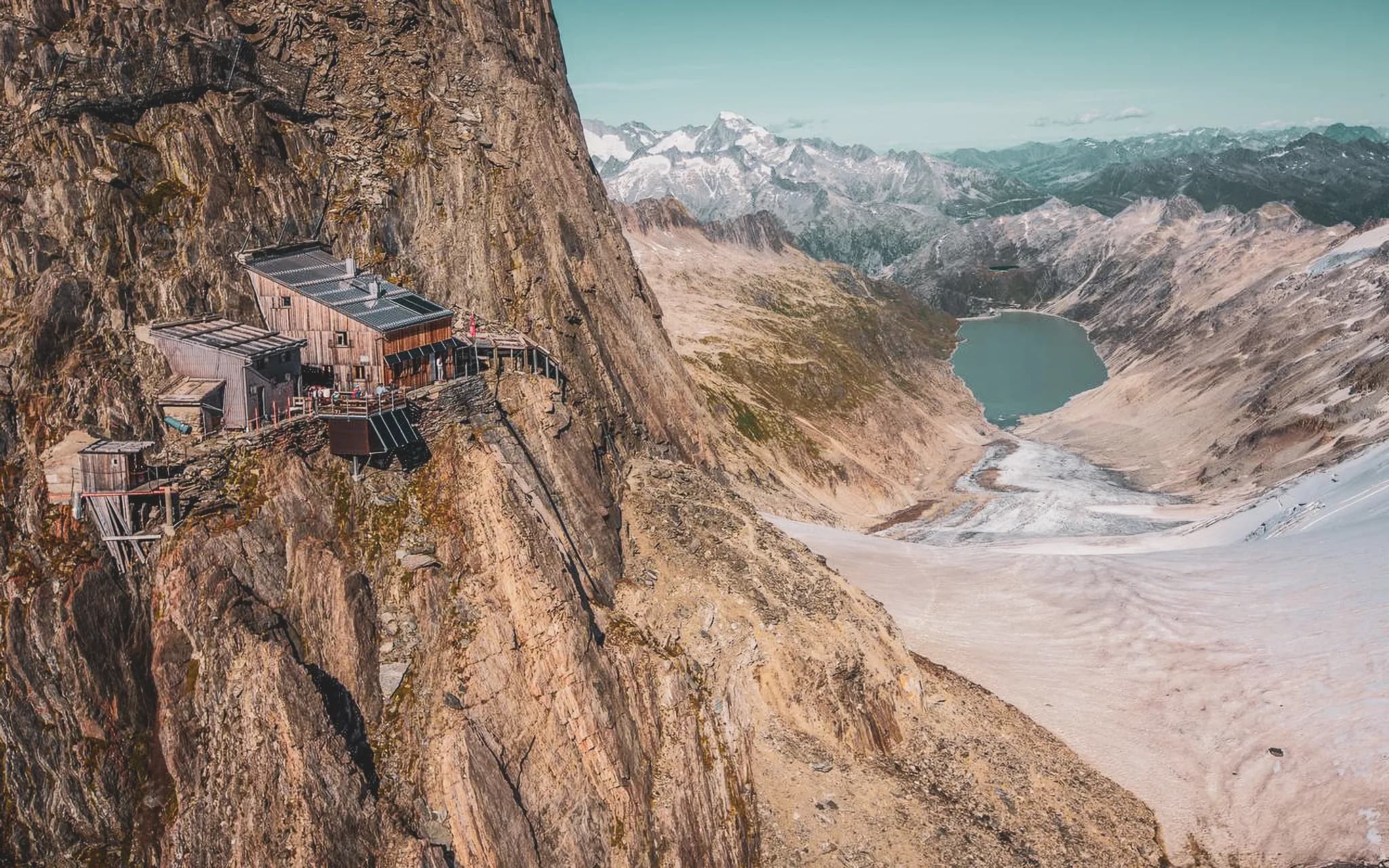 A wooden mountain hut set high on a steep cliff, offering spectacular views over a mountainous landscape. Next to the building, a wooden terrace stretches out, allowing visitors to enjoy the fresh air and panoramic views.