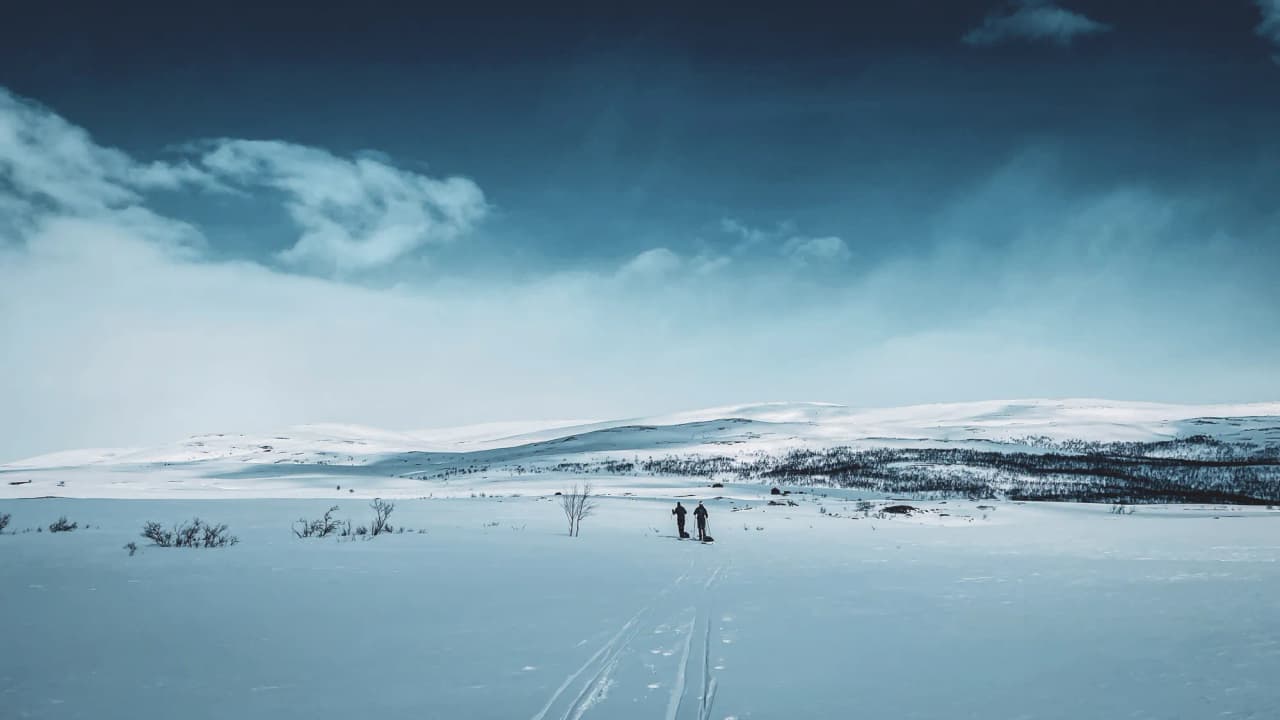 Skiërs doorkruisen sneeuwvrije landschappen onder een blauwe hemel die wordt verlicht door wolken.
