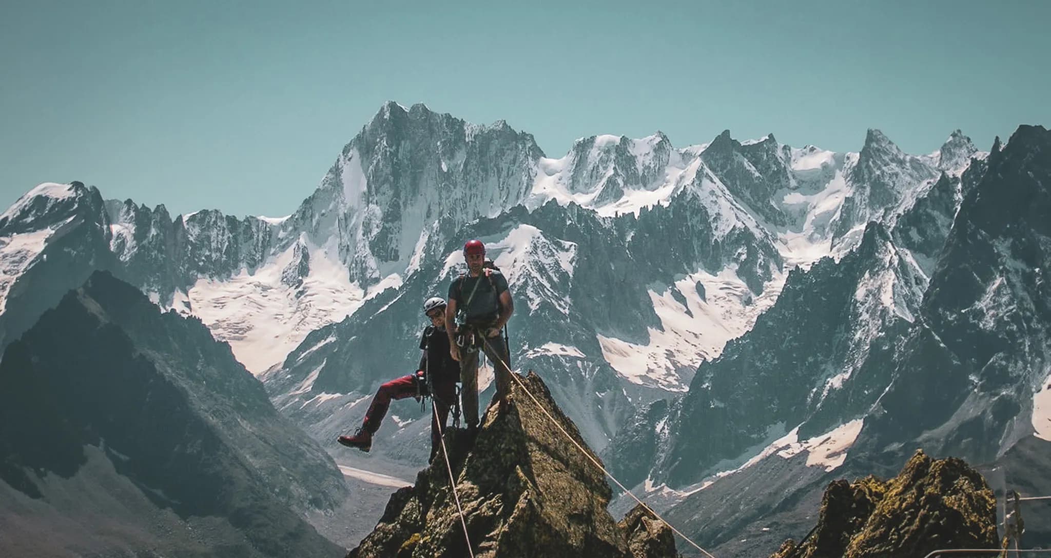 Two climbers are standing on a rocky summit, surrounded by majestic snow-capped mountains. One is seated with one leg bent, while the other is standing, all in the same position.