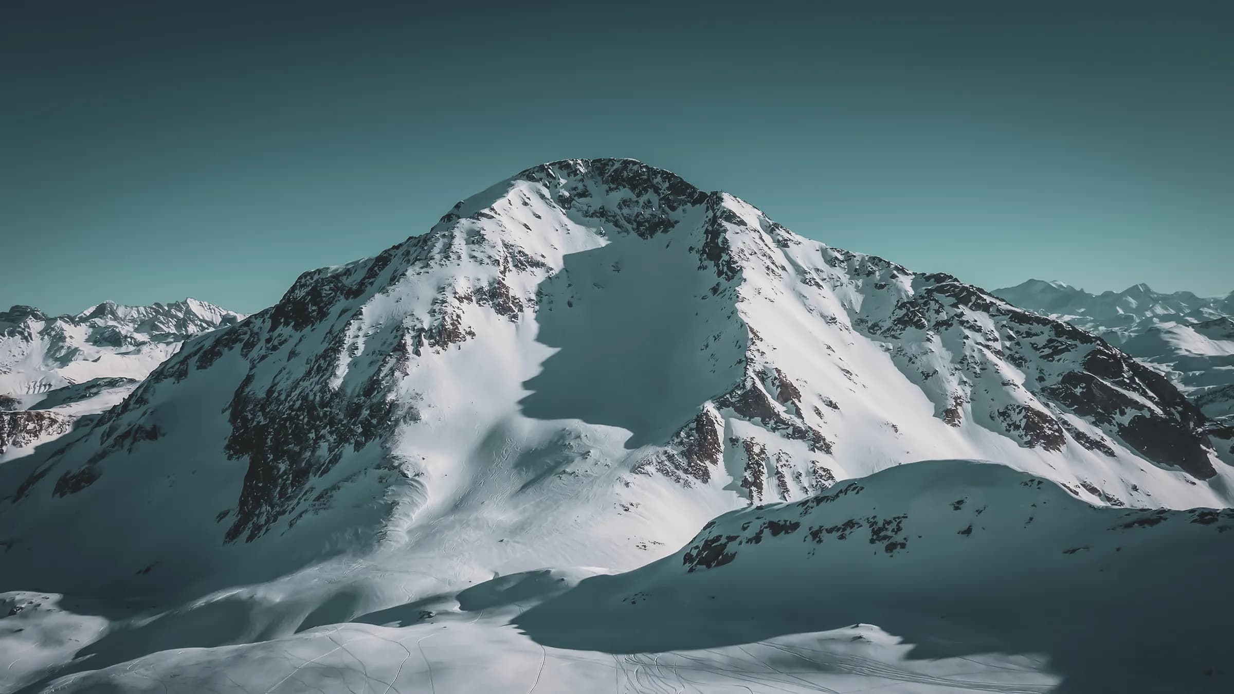 A majestic mountain peak covered in snow, with steep slopes and distinct shadows created by the relief. The sky is a deep blue, providing a clear, serene atmosphere. In the background