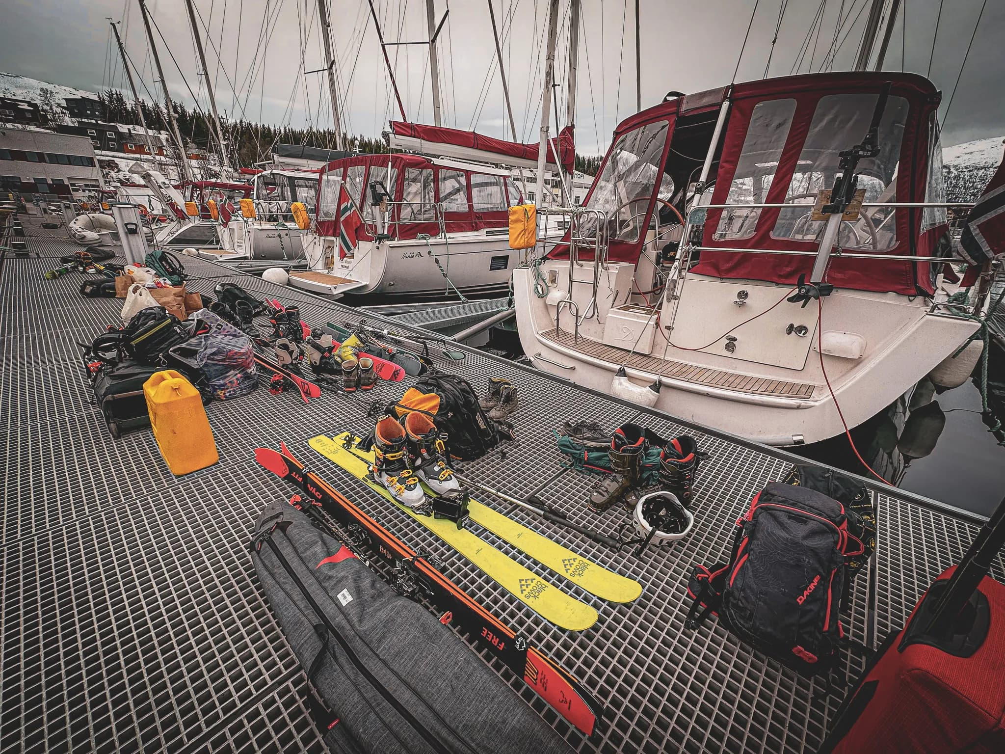Un quai de marina avec plusieurs voiliers amarrés, entouré d'équipements de sports d'hiver. Au premier plan se trouvent des skis jaunes, des chaussures de ski, des sacs à dos et divers accessoires éparpillés sur le