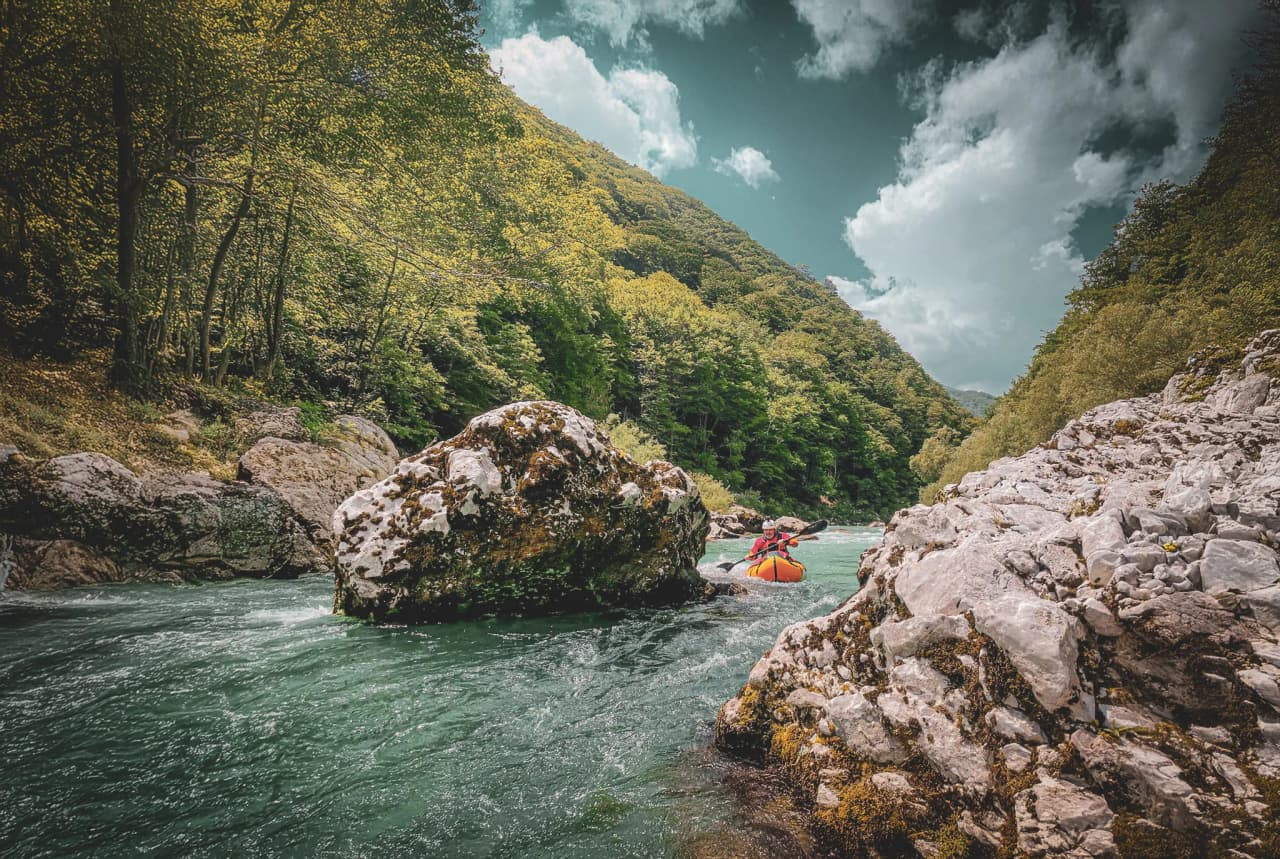 A hiker is paddling a canoe on a fast-flowing river, surrounded by lush green landscapes. Large rocks and boulders litter the banks, while dense, leafy trees stand in the distance.