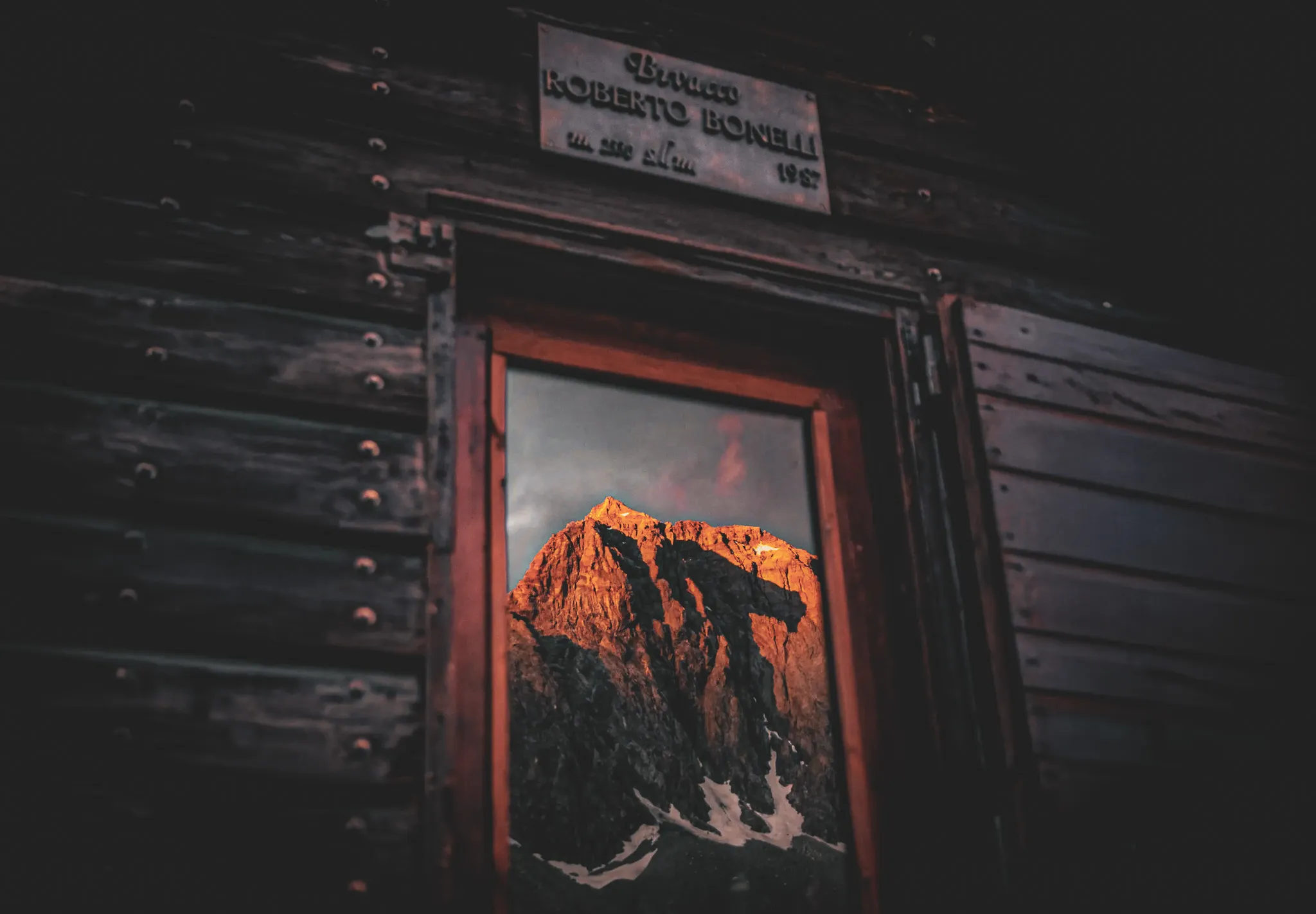 A wooden window through which a mountain landscape is reflected. Outside, craggy peaks illuminated by the golden light of dusk, almost mountainous, contrast with the dark sky. The