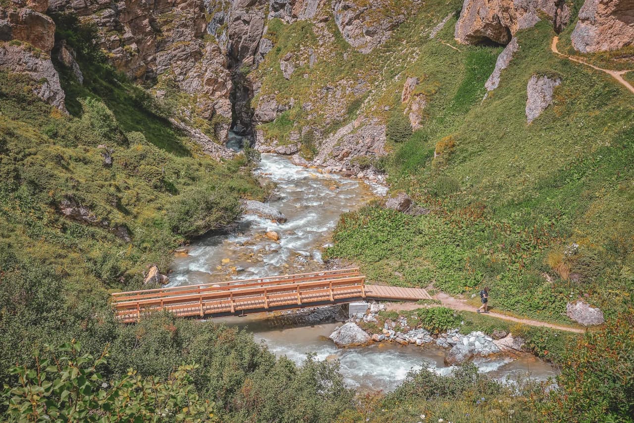 A wooden bridge crosses a lively river, surrounded by greenery and majestic Alpine scenery.