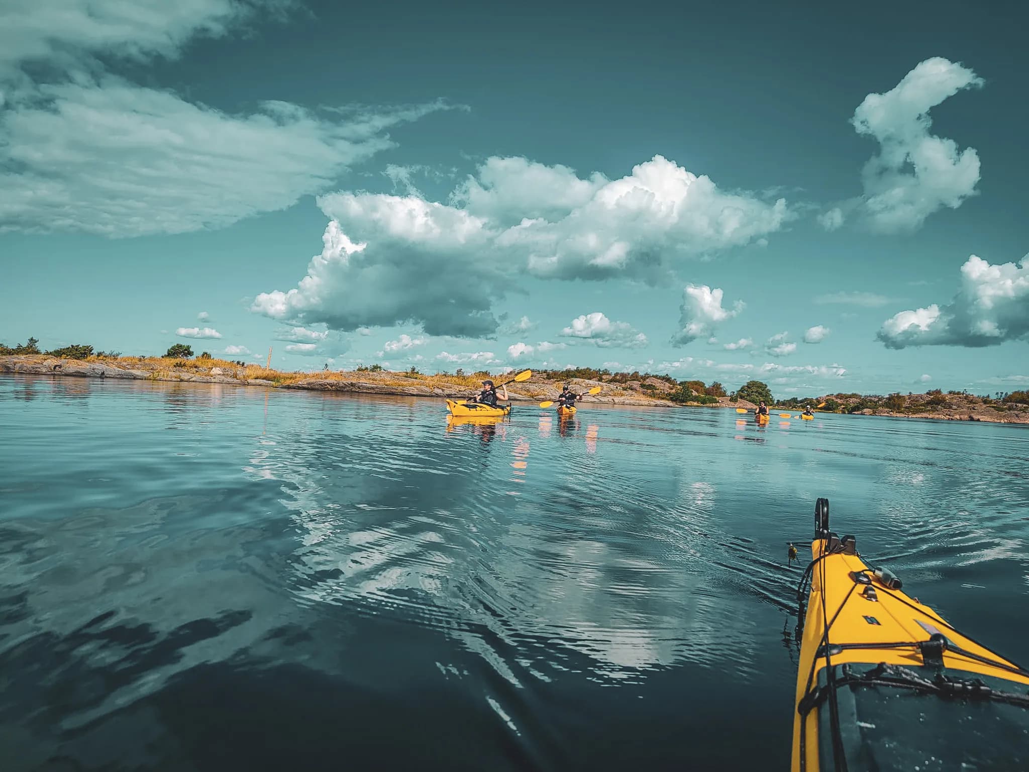 A calm waterscape with several yellow kayaks floating on clear water, reflecting the sky dotted with white clouds. Rocks and lush green vegetation line the shore, adding to the beauty of the water.