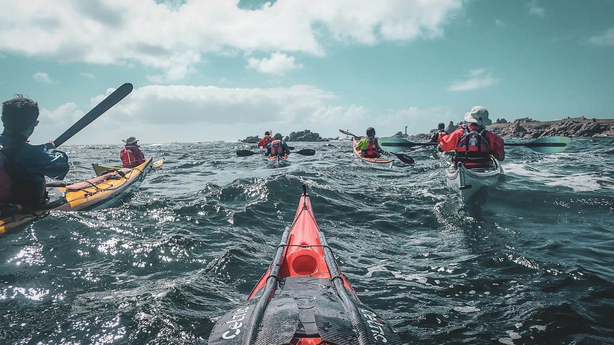 A group of kayakers navigate rough waters, visible from the back of a red kayak in the foreground. The participants are wearing lifejackets and, in some cases, caps or hats.