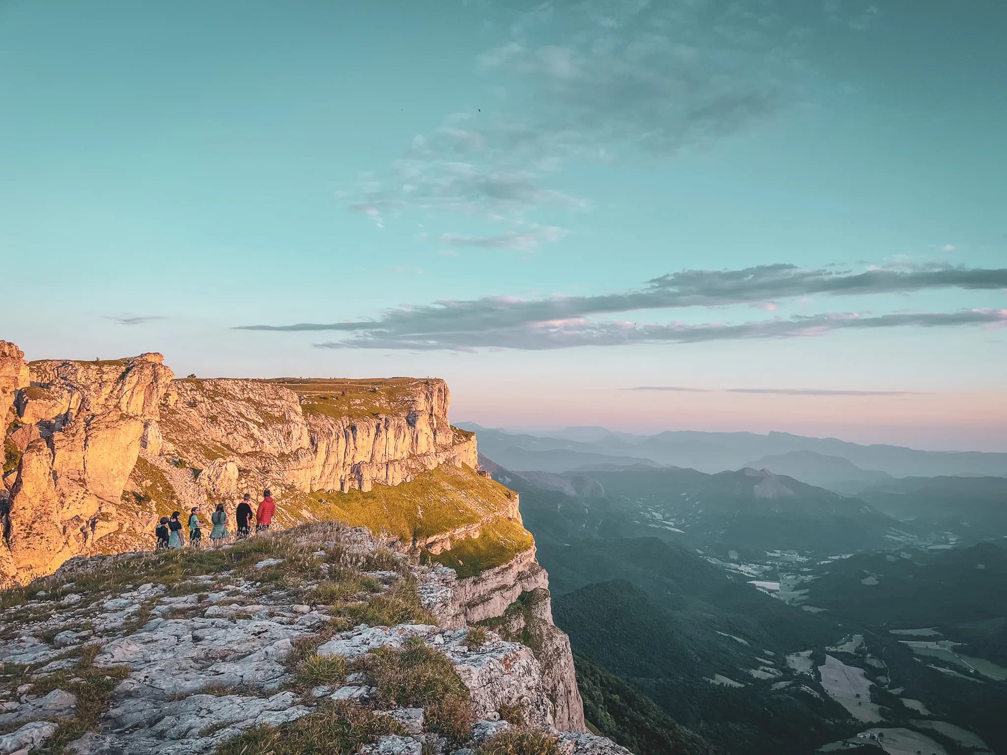 A group of people walk along a rocky ridge, overlooking valleys and mountains in the background. The cliffs feature impressive geological formations, while the sunlight illuminates the paths.