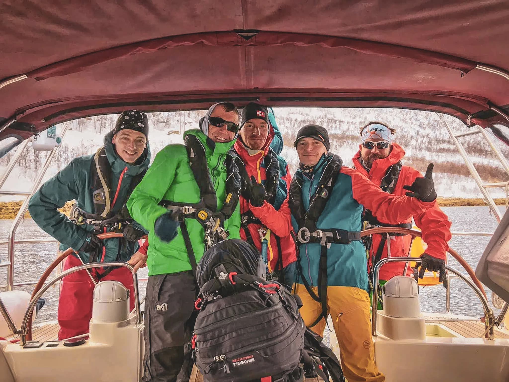 Un groupe de cinq personnes se tient sur un bateau, vêtues d'équipements d'extérieur colorés adaptés pour des activités en milieu aquatique. L'un d'entre eux porte une veste verte, tandis que les autres sont habillés en rouge, bleu