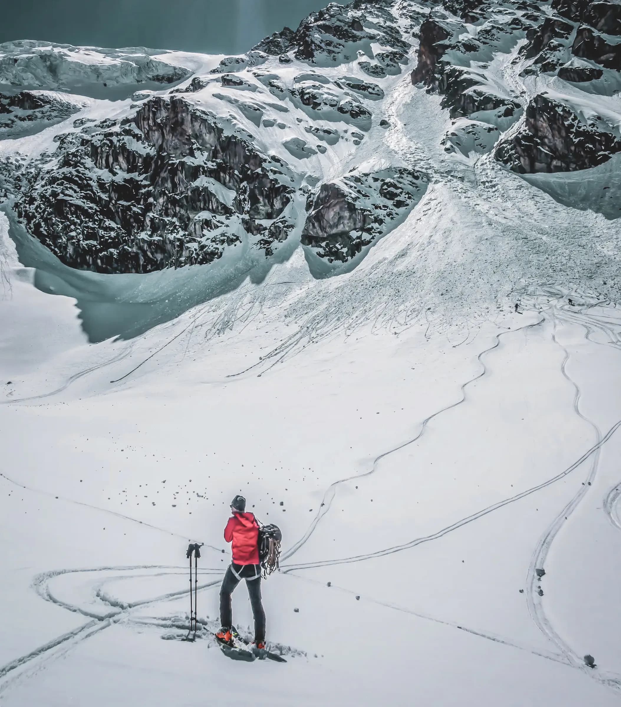 A skier stands on a vast expanse of snow, gazing up at an imposing range of snow-capped mountains under a luminous sky. The sun is shining brightly, creating a striking contrast with the landscape. The snow is jon