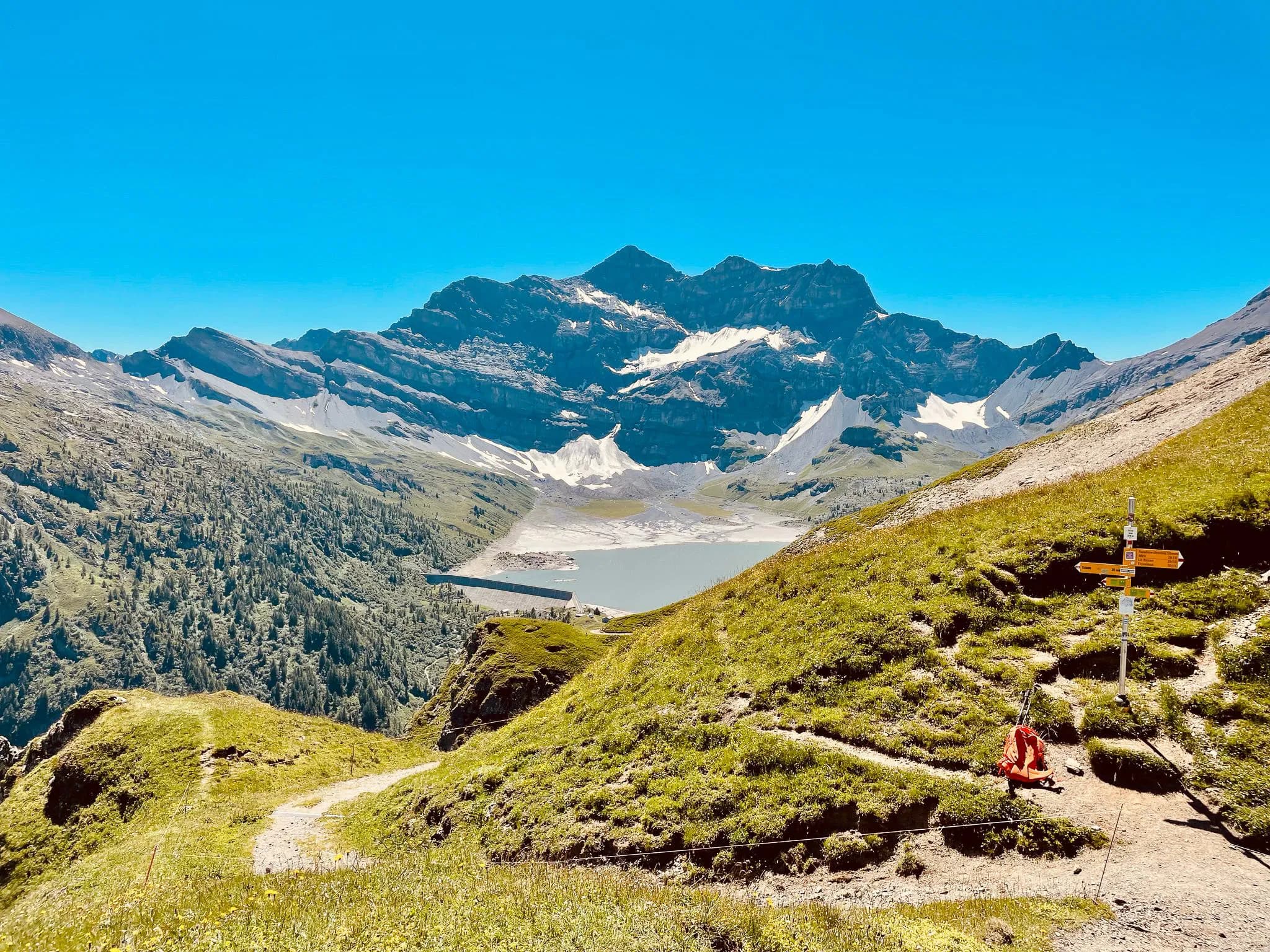 An impressive mountain landscape with majestic peaks and glaciers visible from the summit. In the foreground, a winding path crosses a green meadow dotted with wild flowers. To the right, a signpost