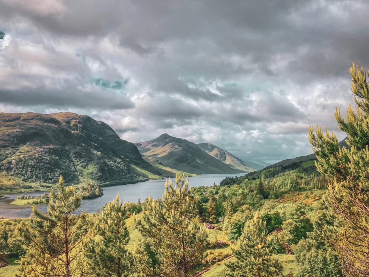 Panoramic view of the Scottish Highlands, lush green valleys and sparkling lochs under cloudy skies.