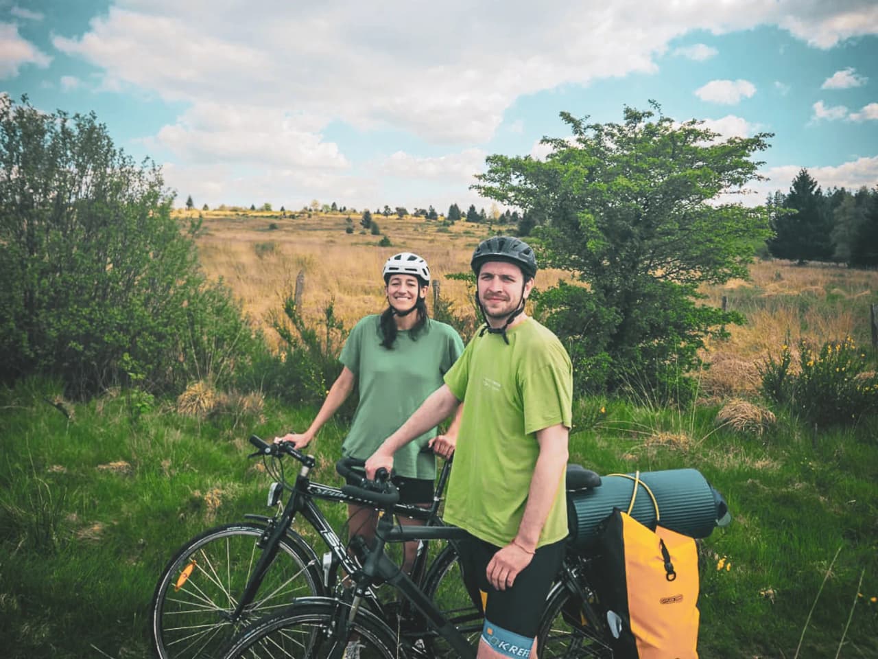 Deux cyclistes souriants dans les Hautes Fagnes, entourés de verdure, prêts pour l'aventure.
