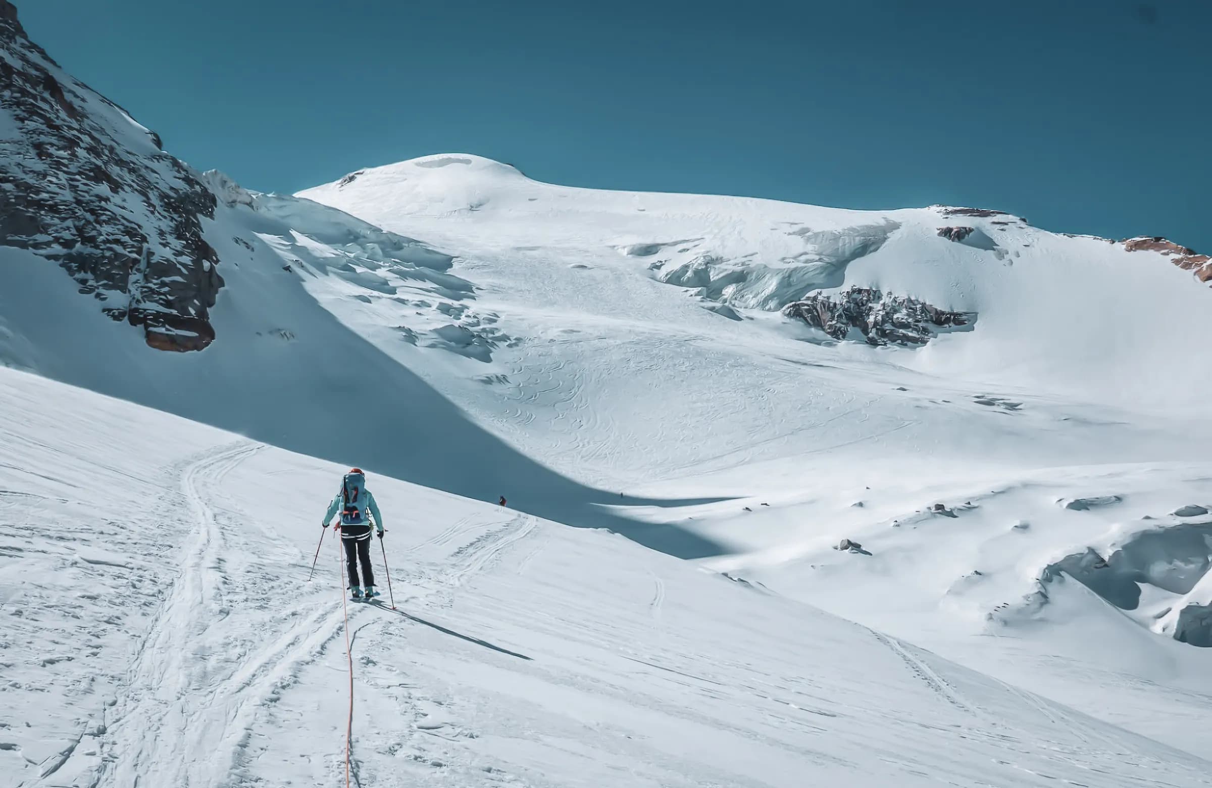 A skier in full effort on a snow-covered slope, surrounded by majestic snow-capped mountains. The sky is a brilliant blue, contrasting with the immaculate white of the landscape. Tracks