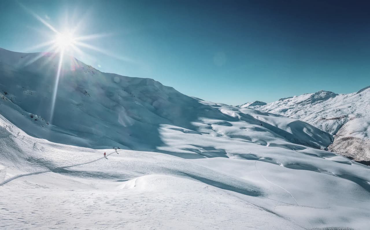 A snowy landscape bathed in light, with gentle mountains and immaculate slopes beneath a clear blue sky. The sun shines intensely, creating flashes of light on the snow, while a silhouette