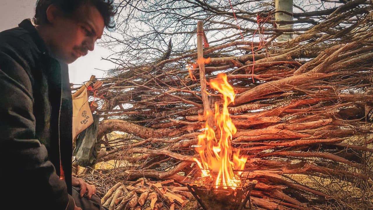 A man with curly hair, meditating by a campfire, surrounded by branches, immersive.