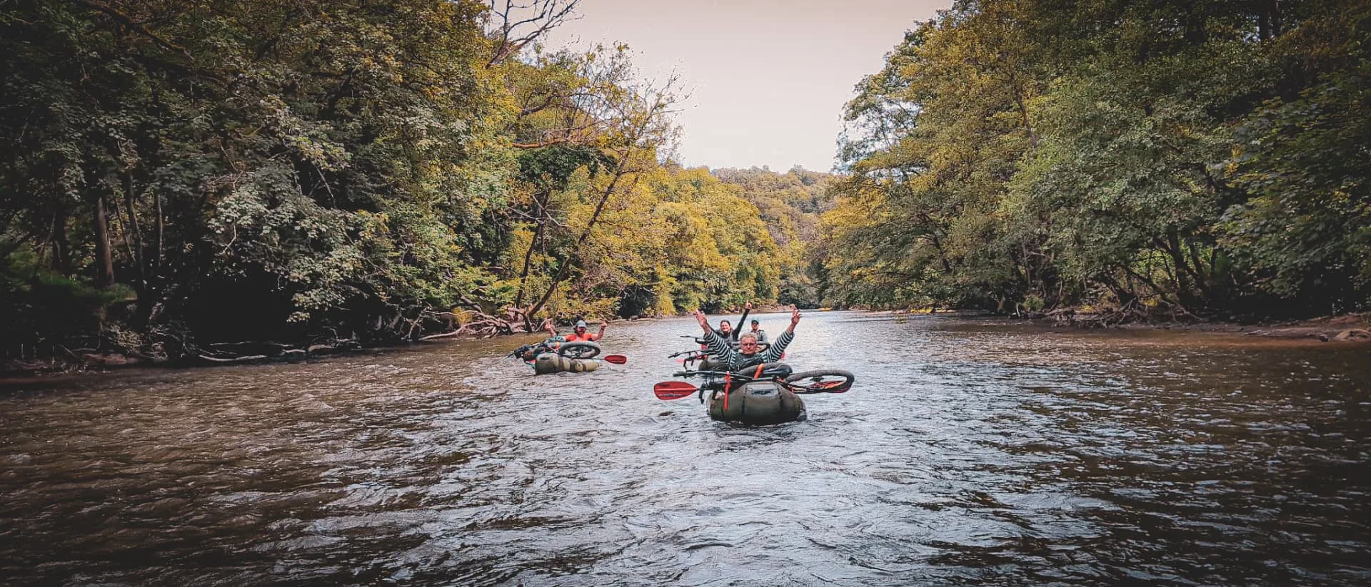 A group of people are tubing on a river surrounded by dense vegetation. The participants, in buoys and carrying paddles, seem to be enjoying themselves and some are raising their arms in joy. Coloured trees