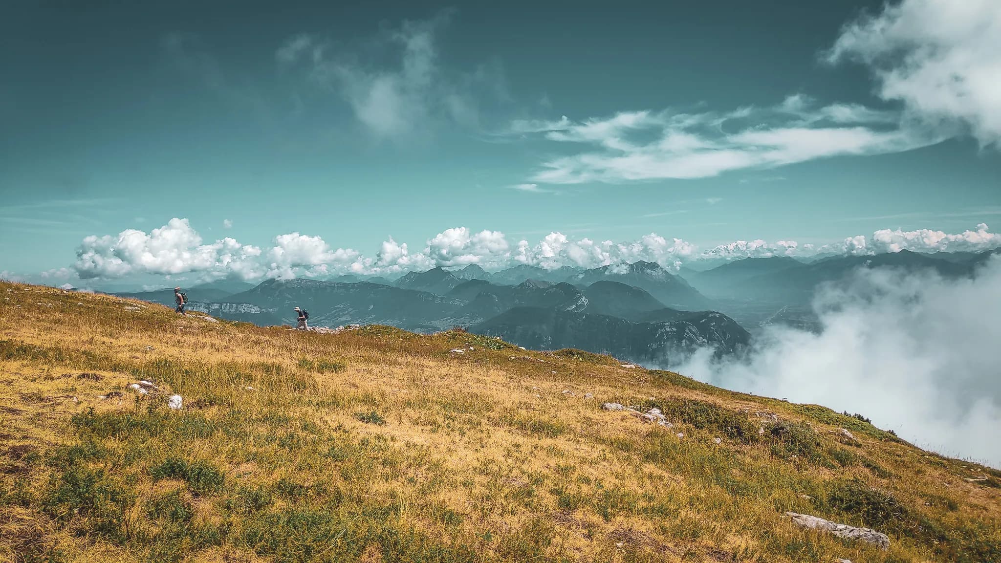 Un paysage montagneux s'étend à perte de vue, avec des prairies verdoyantes au premier plan. Deux personnes marchent sur le terrain, profitant de la vue panoramique. En arrière-plan, des montagnes majestueuses se