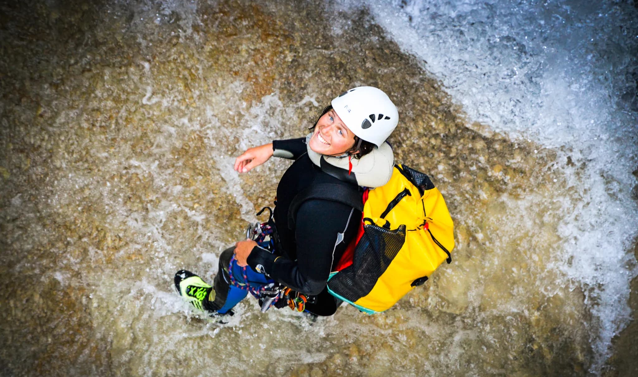 A smiling person is standing in the water, wearing a white helmet and a black wetsuit. She is carrying a large yellow rucksack and is surrounded by small splashes of water and rocks. The background appears to be a