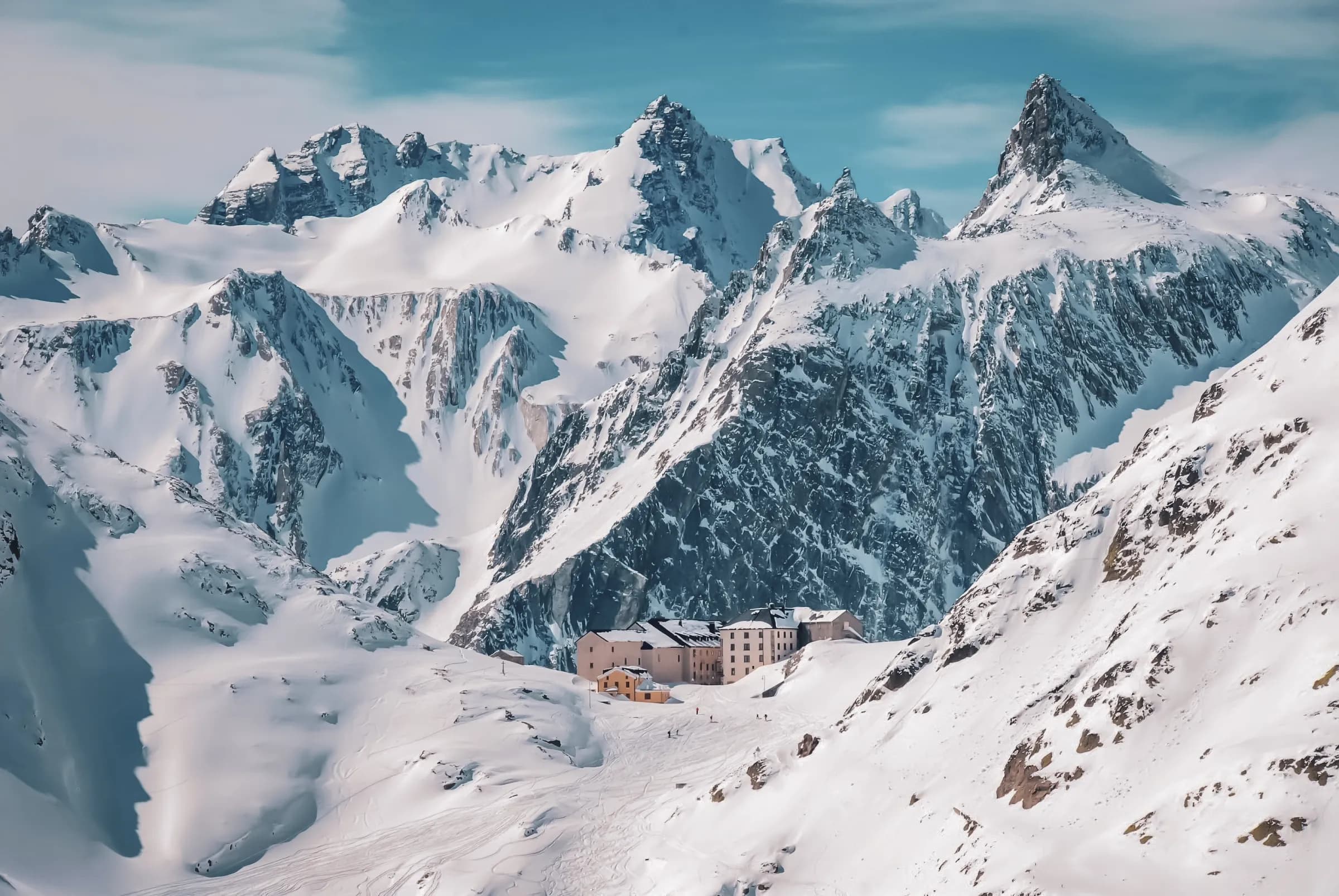 A spectacular mountain landscape, with majestic snow-capped peaks rising up under a clear blue sky. Below, an Alpine-style building is visible, probably a refuge or hotel, surrounded by fresh snow. The