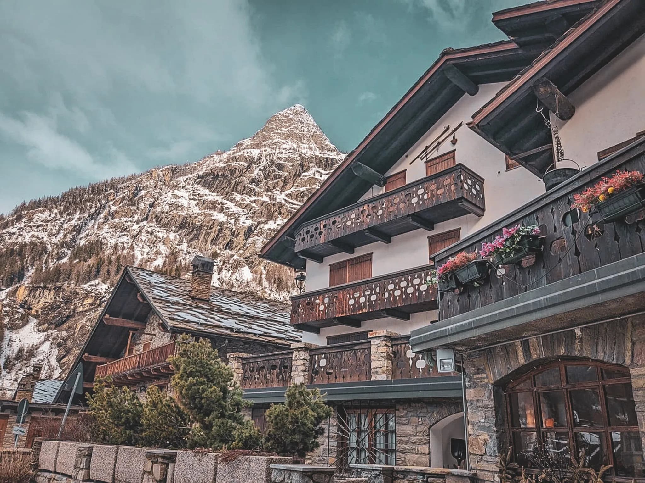 Un chalet en montagne, construit en pierre et en bois, avec un toit en ardoise. La structure présente des balcons ornés de balustrades sculptées et de fleurs en pots. En arrière-plan, un paysage montagneux se d