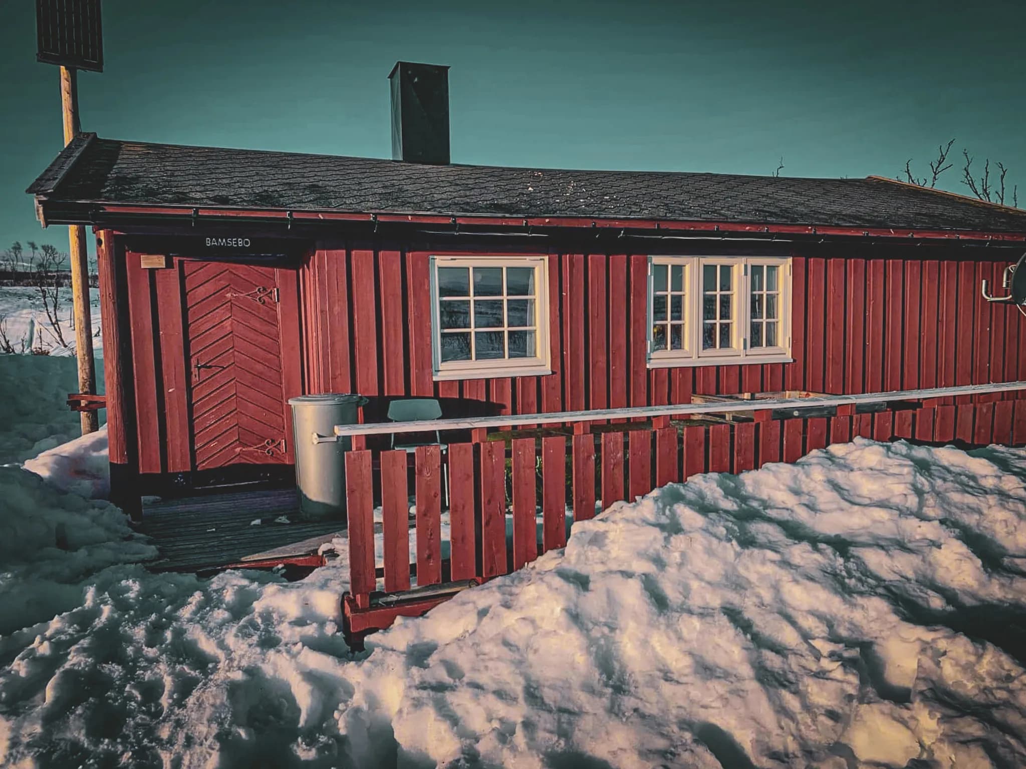A red wooden chalet is surrounded by snow. The structure has a wooden door with diagonal panels, several white windows and a small wooden balcony. Next to the entrance is a rubbish bin, and a