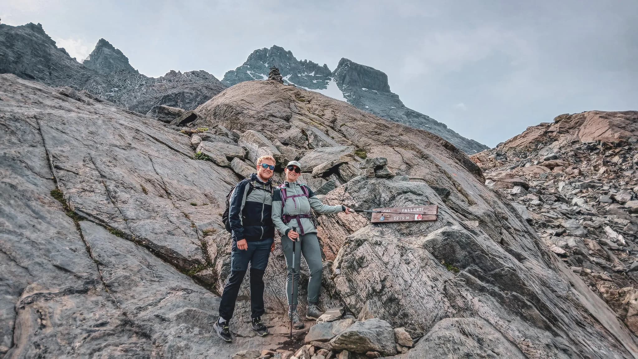 Twee wandelaars poseren op een alpenpad, omringd door majestueuze rotsen en bergtoppen.