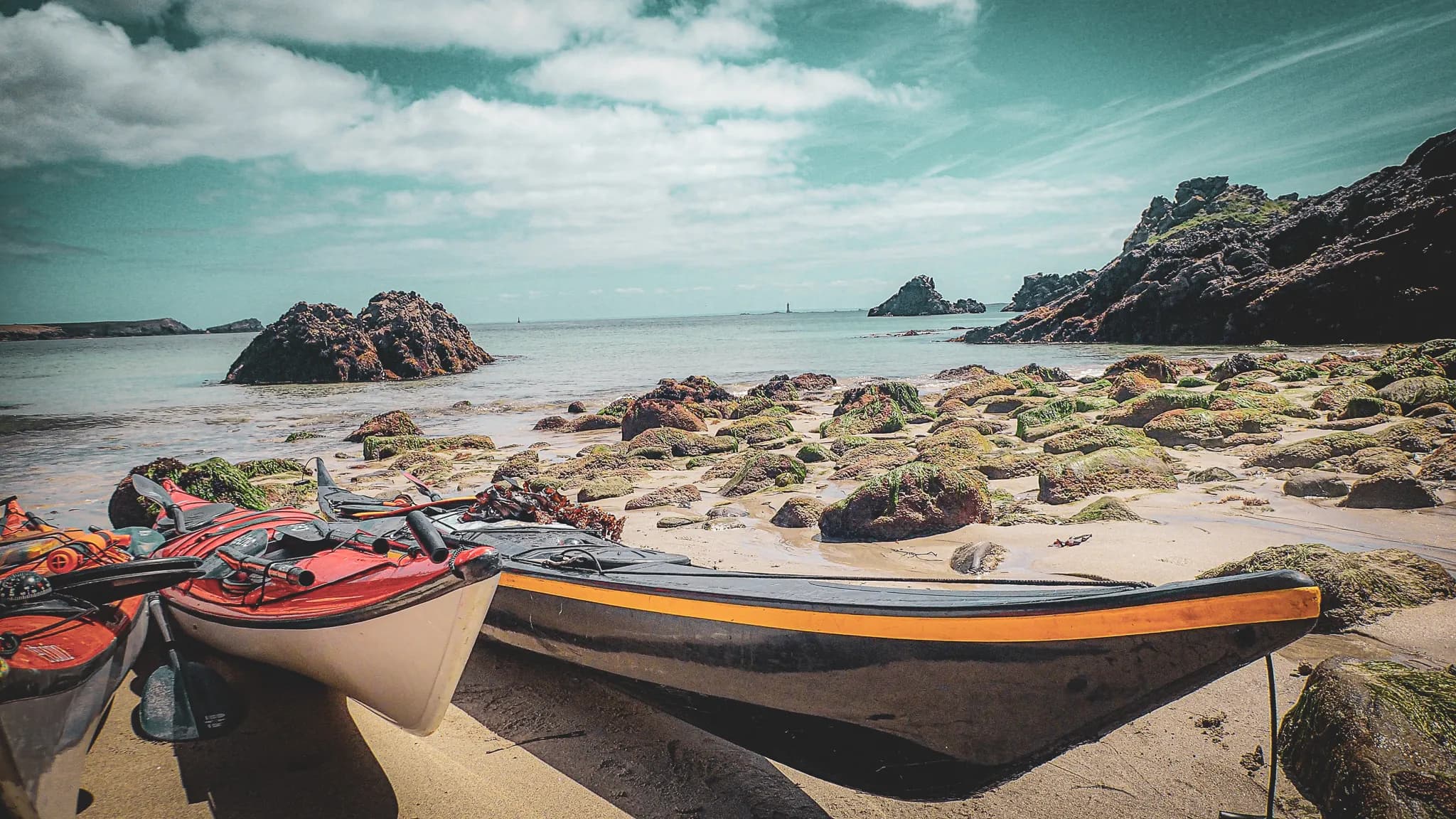A sunny shoreline with kayaks on a sandy beach, surrounded by seaweed-covered rocks. The clear, calm water reflects the cloudy sky. In the background, rocky mountains