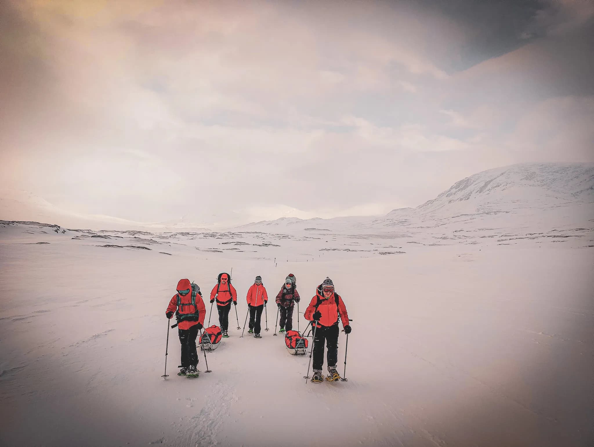 A group of people on a snowshoe hike in snowy terrain, dressed in brightly coloured winter clothing, including red jackets. Some are pulling pulkas, while others are using walking sticks.