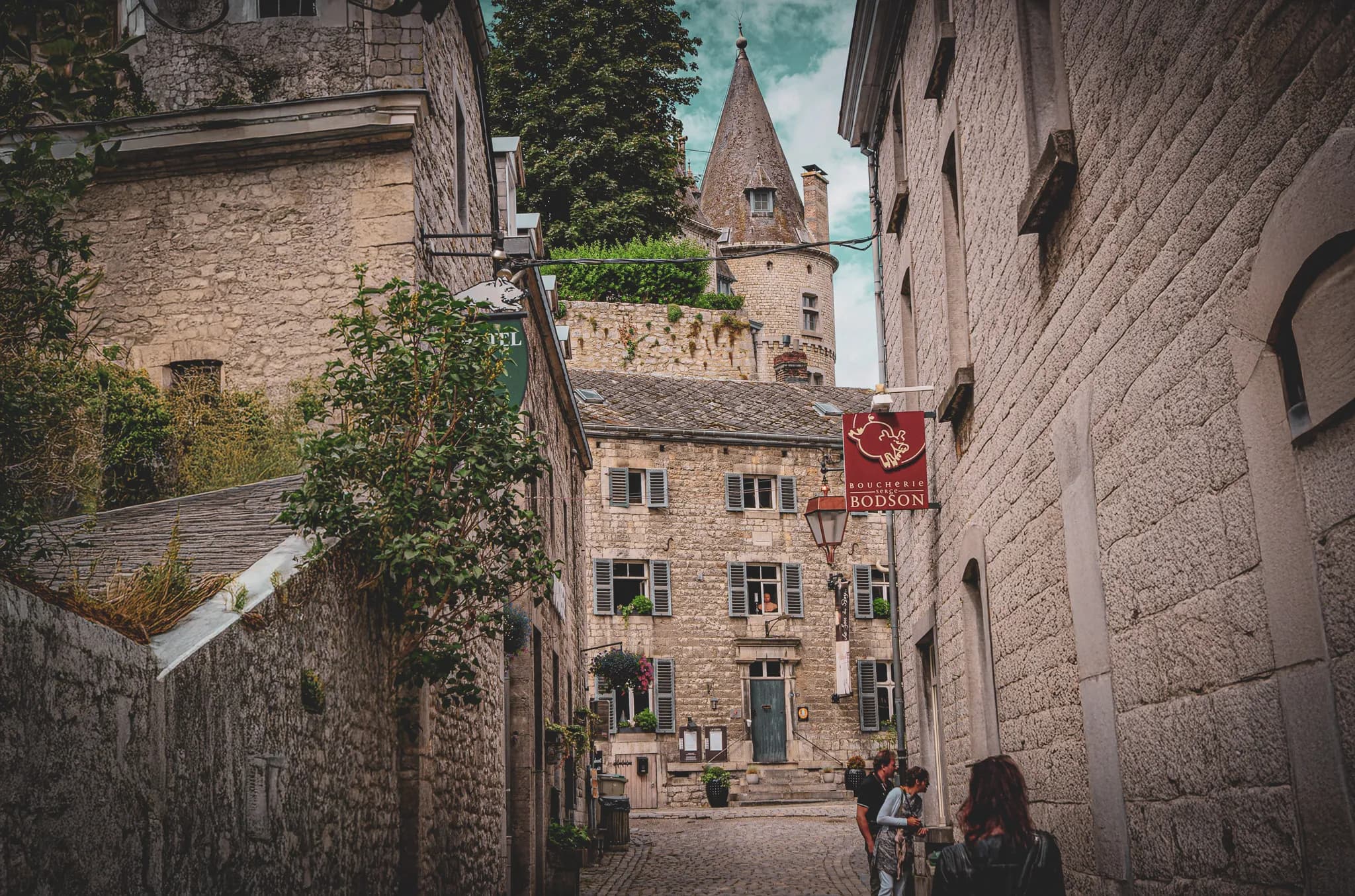 A cobbled lane lined with stone houses, with blue shutters and ornamental plants. At the far end, a turreted building dominates, while a red sign indicates a butcher's shop. Pass