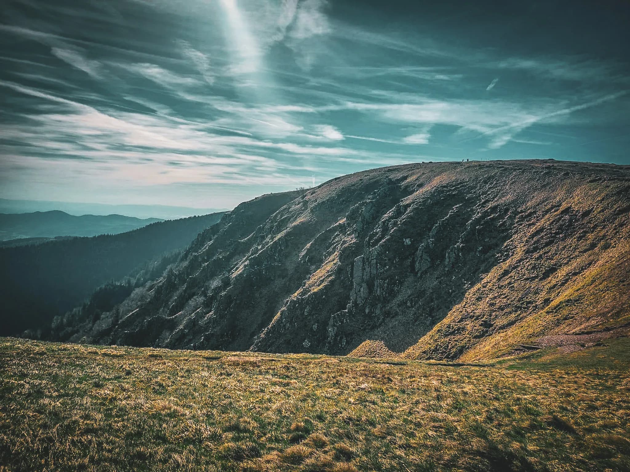 A vast mountain landscape with grassy and rocky slopes, stretching out under a partly cloudy sky. Shades of green and brown dominate the ground, while silhouettes of walkers can be seen on the horizon.