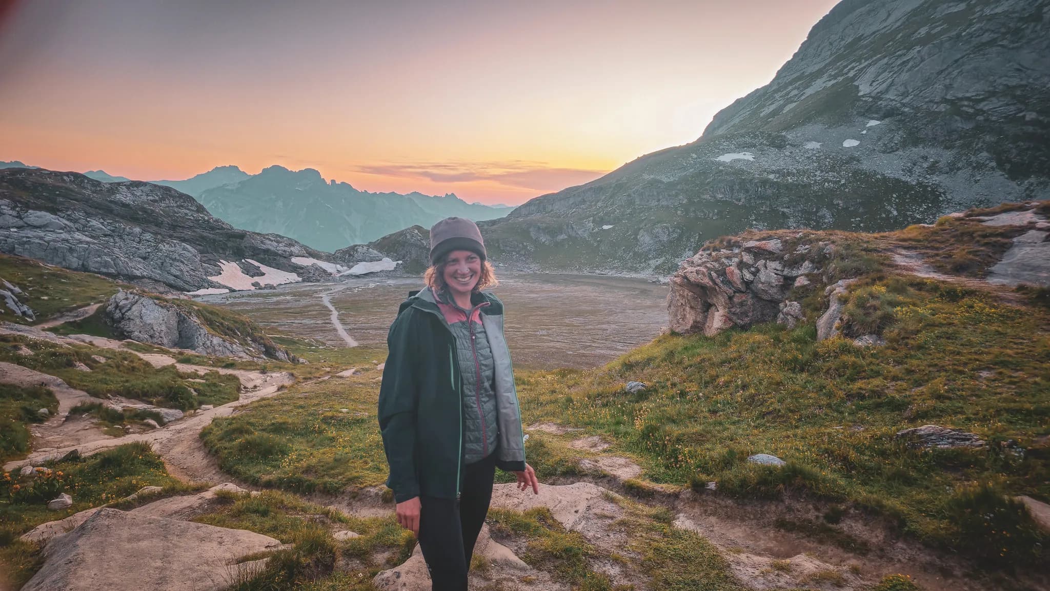 Un paysage alpin au coucher du soleil, avec des montagnes majestueuses en arrière-plan. Une personne souriante se tient sur un sentier de randonnée entouré de verdure, portant une veste et un bonnet, évoquant une atmosphère d