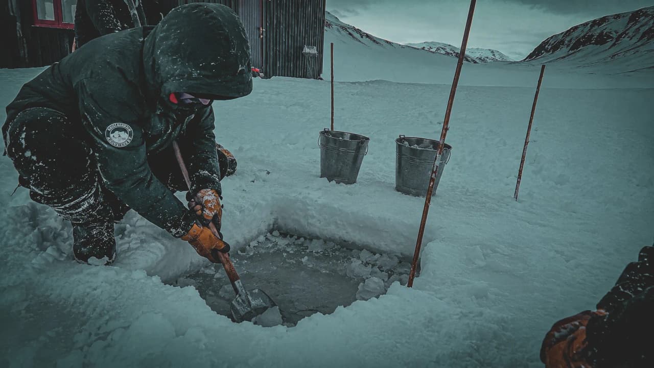 Een avonturier graaft zich een weg door de sneeuw om bij gletsjerwater te komen, omringd door besneeuwde landschappen.