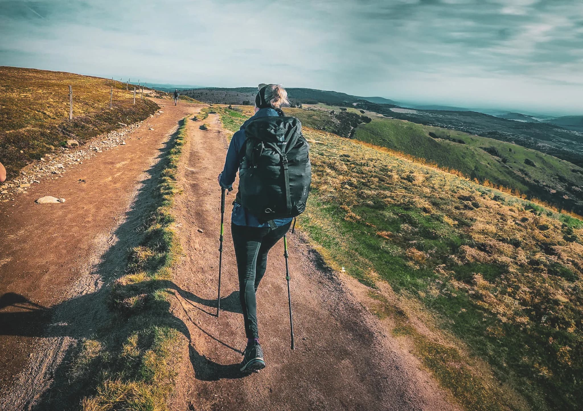 A dirt path bordered by greenery winds through a hilly landscape. One person is walking with his back turned, carrying a large rucksack and using hiking poles. The sky is partly cloudy