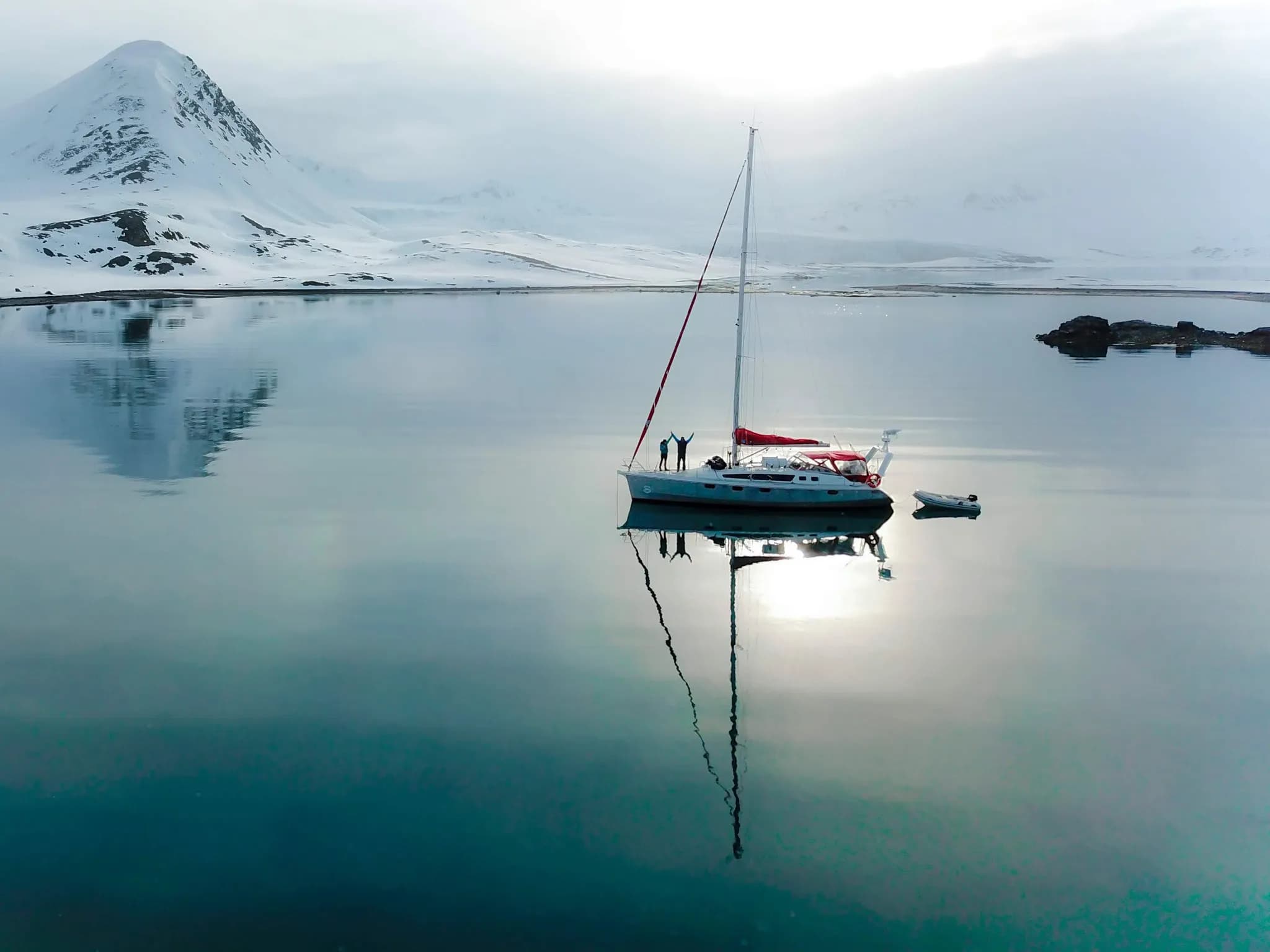 Un voilier navigue sur des eaux calmes entourées de montagnes enneigées. Le ciel est nuageux, créant une ambiance sereine, et la lumière se reflète doucement sur la surface de l'eau. À bord, deux