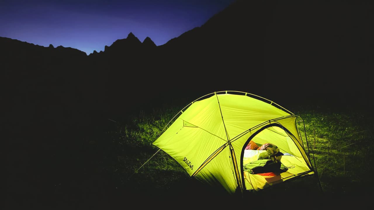 Tente lumineuse sous un ciel étoilé, entourée de montagnes majestueuses. Évasion alpine promise.