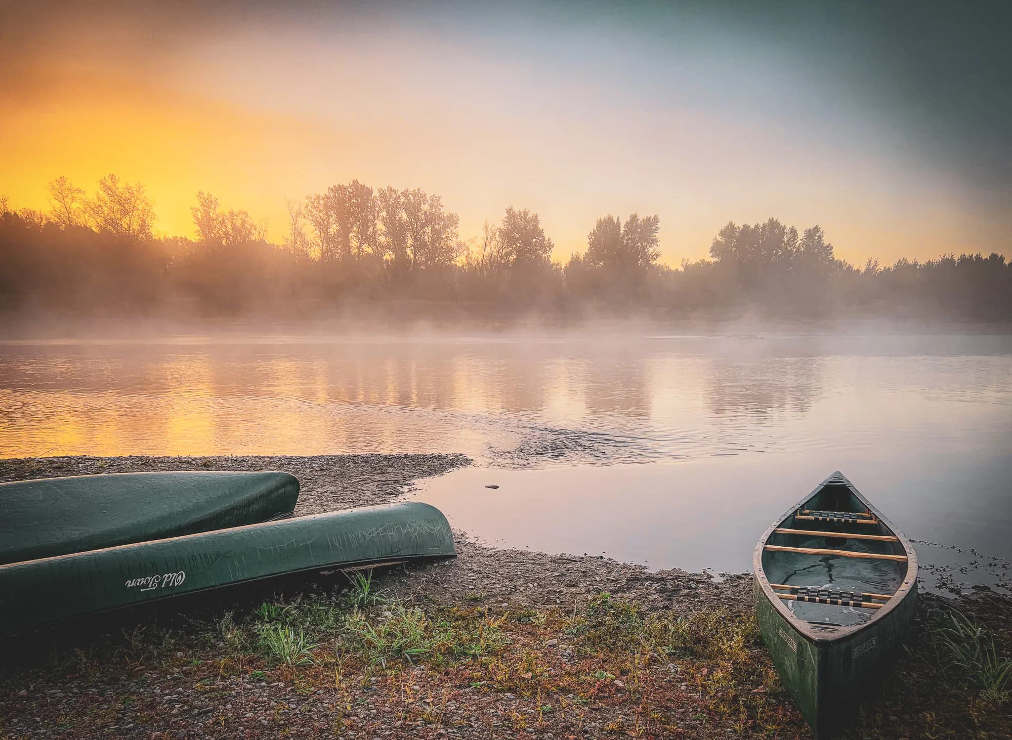 A landscape at sunrise on the banks of a river, with a mixture of light fog and flashes of orange and yellow in the sky. Two green canoes rest on a lightly sandy shore, amid the vegetation of the river.