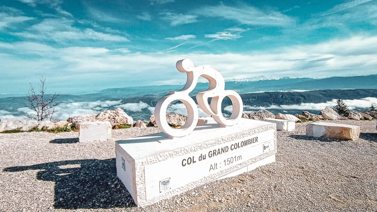 Stylised sign in the shape of a cyclist on the Col du Grand Colombier, with a panoramic view of the Jura massif.