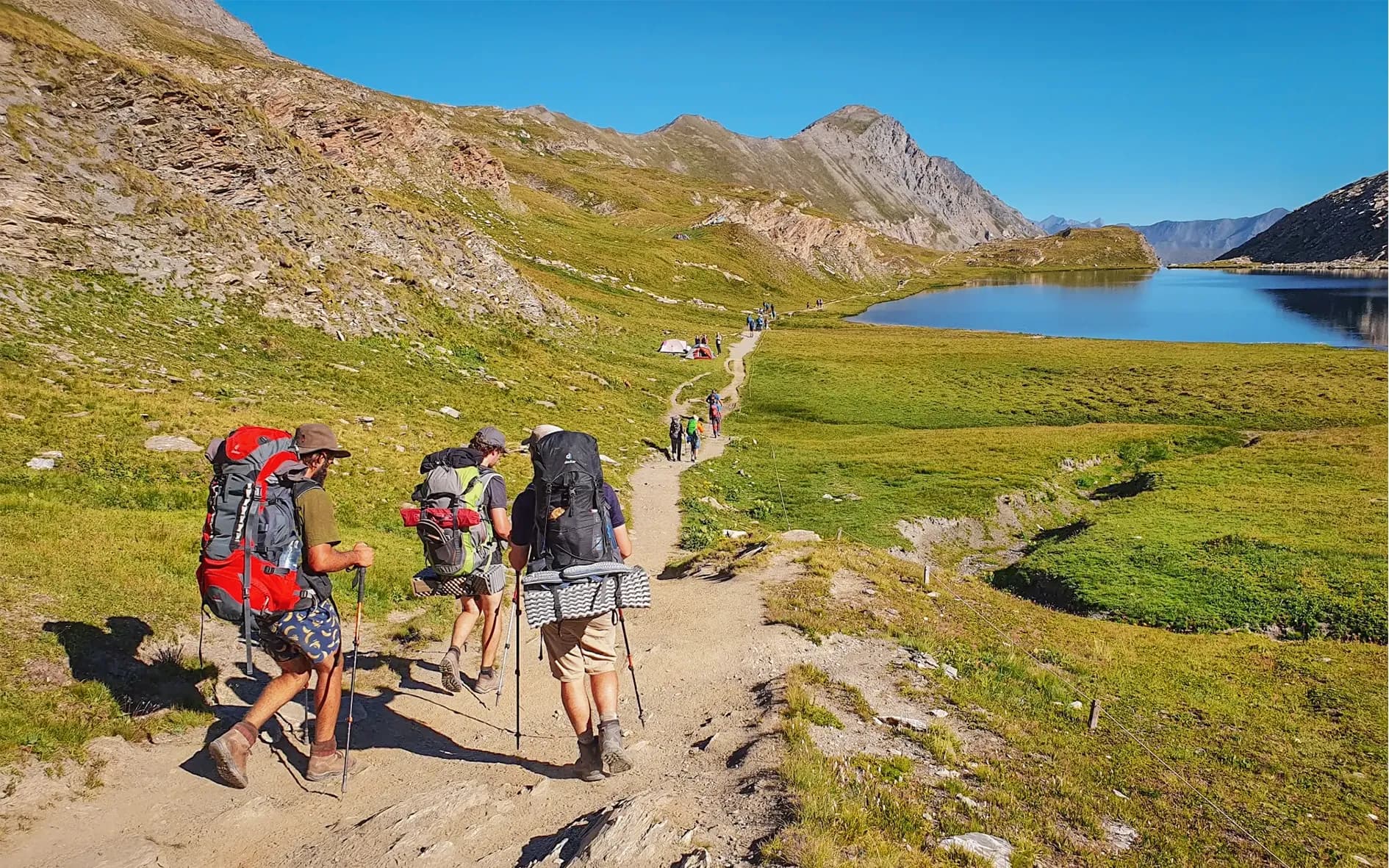 A group of hikers walk along a mountain path surrounded by green meadows and hills. They are carrying large rucksacks and using hiking poles. In the background