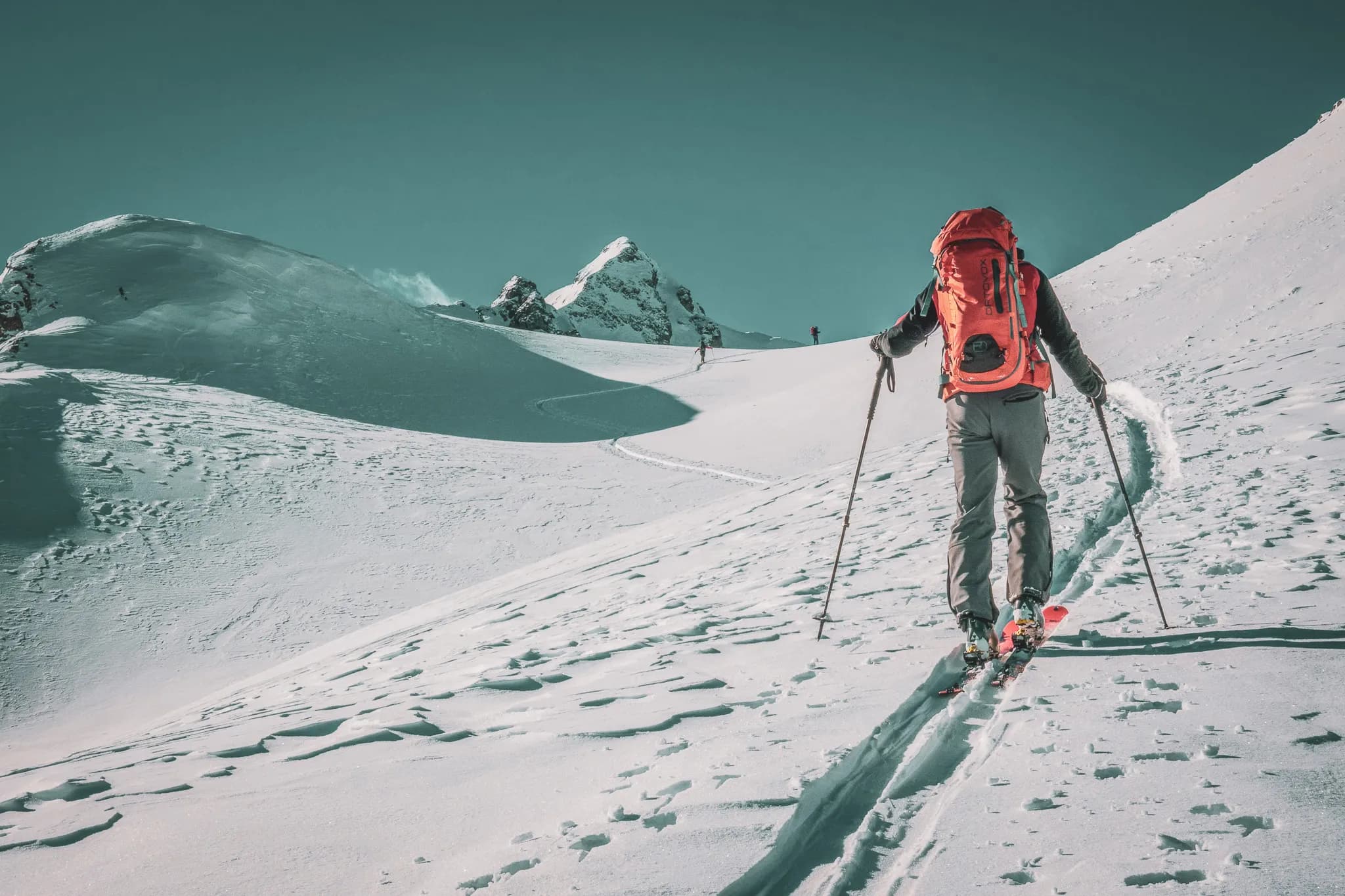 Un skieur traverse un paysage enneigé, portant un grand sac à dos orange et utilisant des bâtons de ski pour se frayer un chemin. Le terrain est vallonné, jonché de traces de pas et de traces de ski. À
