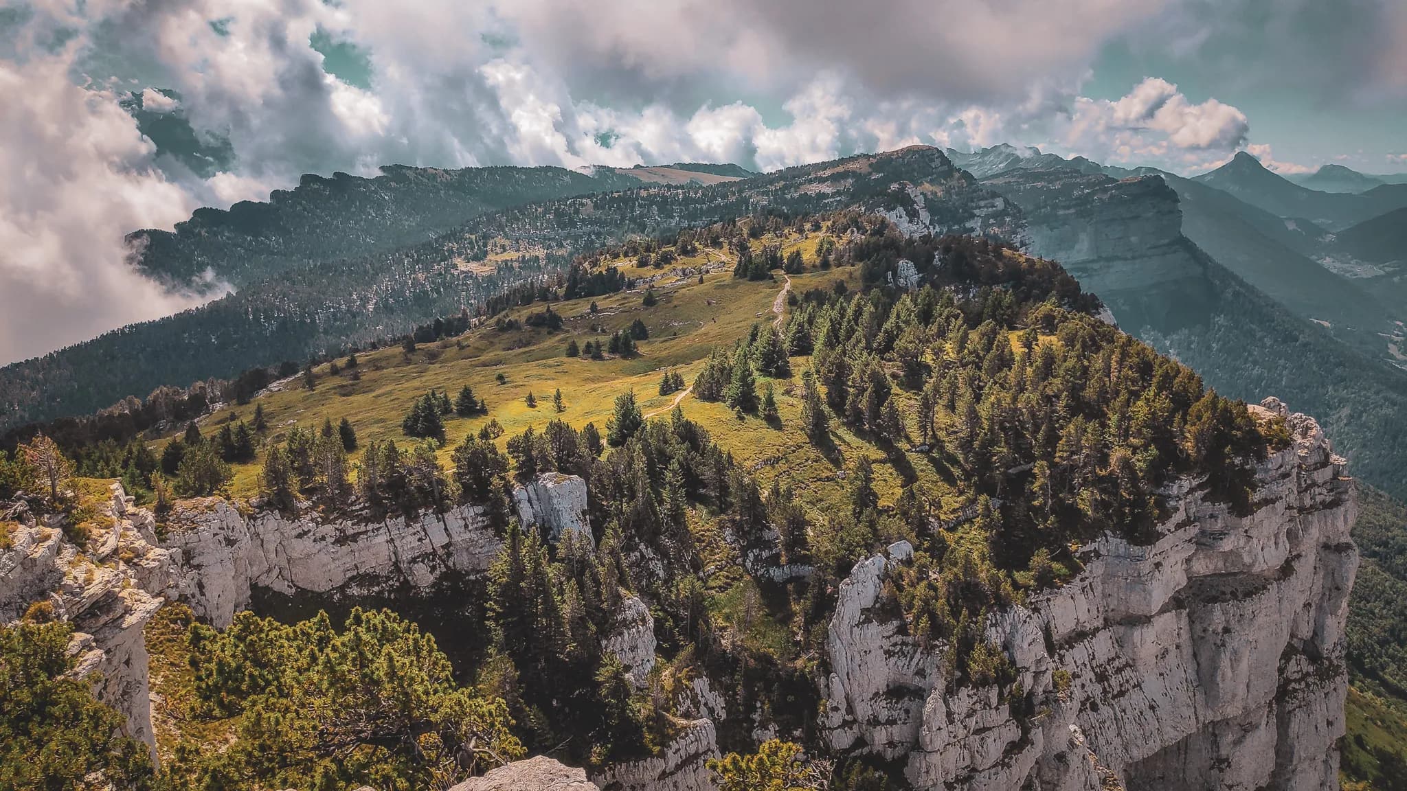 Un paysage montagneux spectaculaire, avec des collines verdoyantes parsemées de conifères, surplombant des falaises rocheuses blanches. Des nuages majestueux flottent dans le ciel, ajoutant une atmosphère dramat