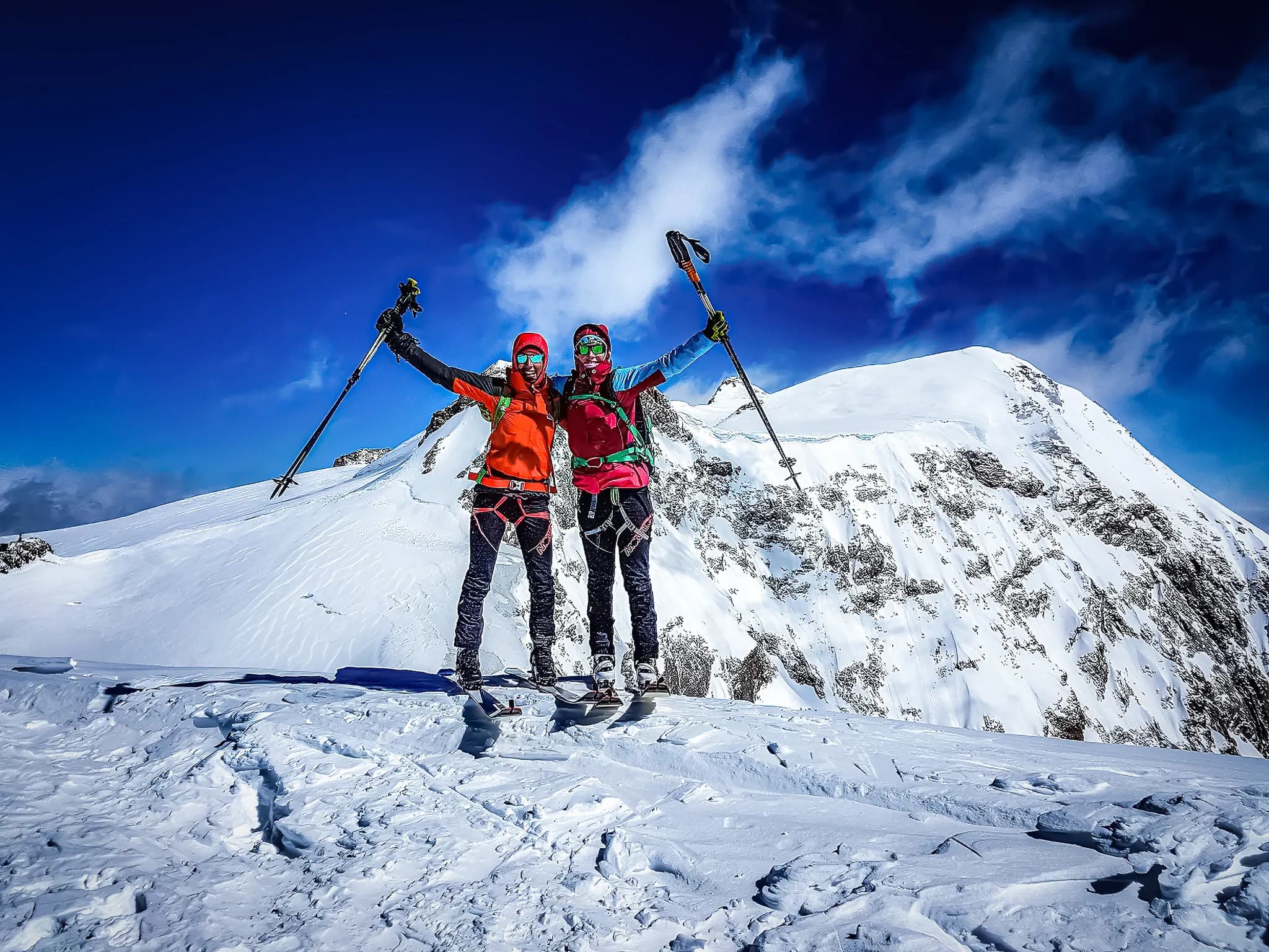 Twee mensen staan op een besneeuwde top, omringd door een majestueus berglandschap. Ze dragen kleurrijke skikleding en uitrusting zoals wandelstokken. Hun gezichten stralen van vreugde,