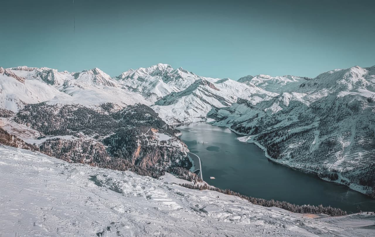 A snow-covered mountain panorama, with majestic peaks under a clear sky. In the centre, a calm lake reflects the surrounding mountains, while coniferous forests line the horizon.